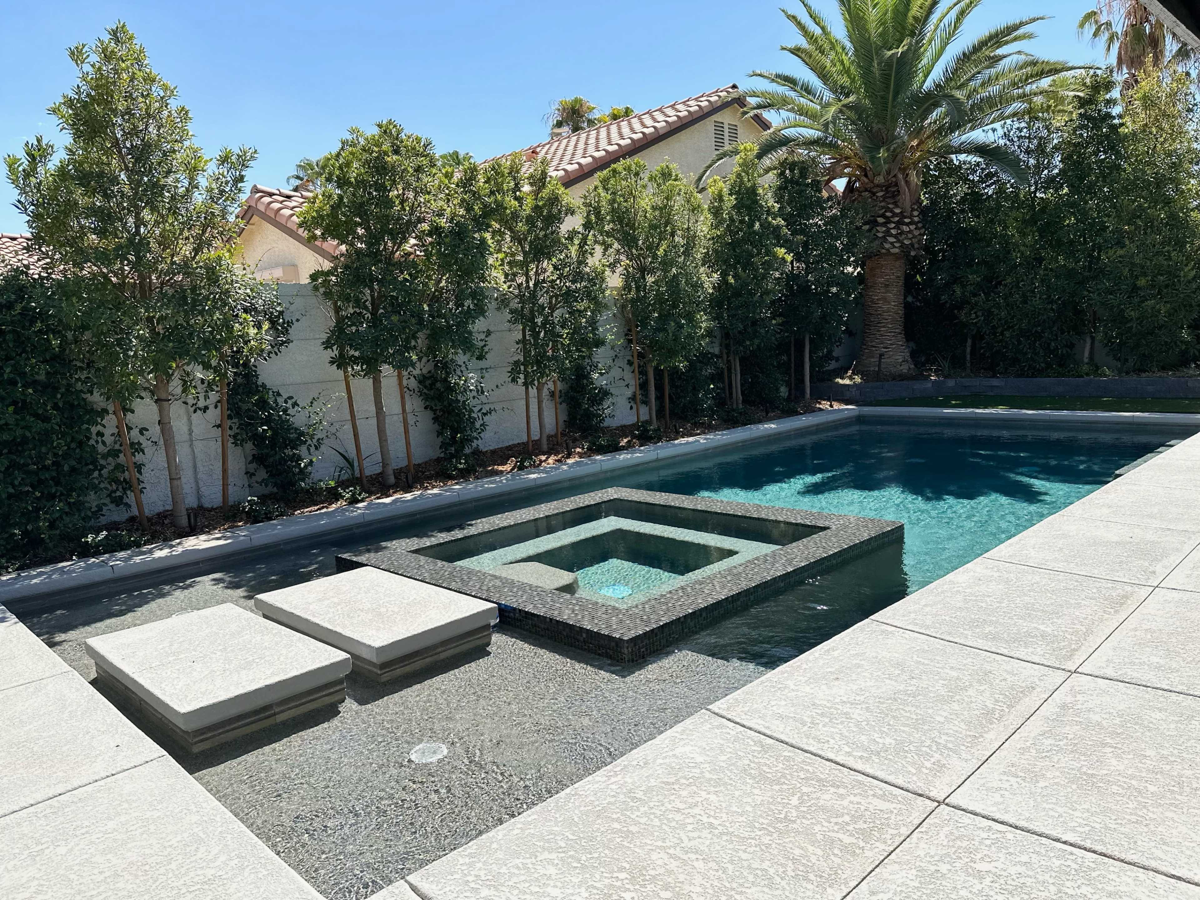 A modern swimming pool featuring a central spa with raised concrete platforms, surrounded by lush greenery and palm trees.