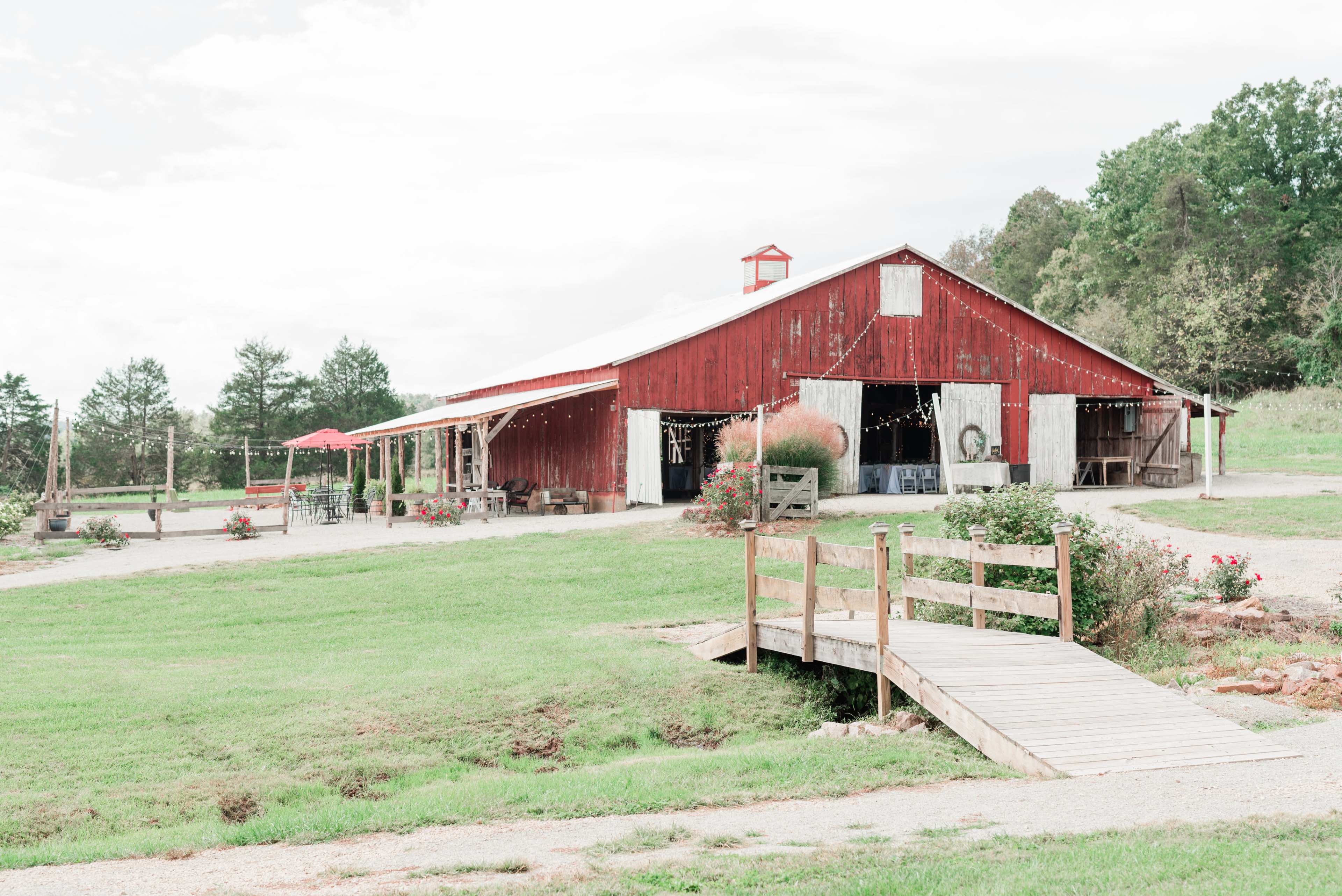 A red barn with a porch and a wooden bridge is situated on a grassy area surrounded by trees.