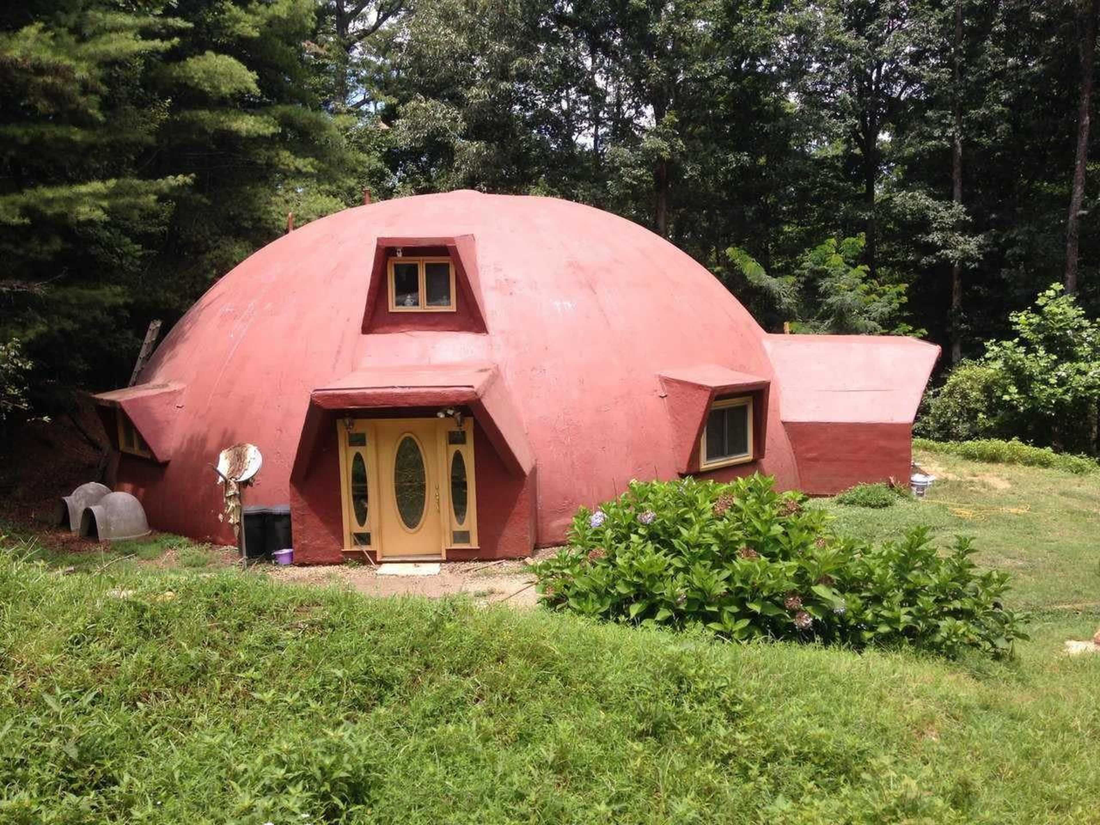 The image shows a red, dome-shaped house with a unique architectural design, set in a green, wooded area.