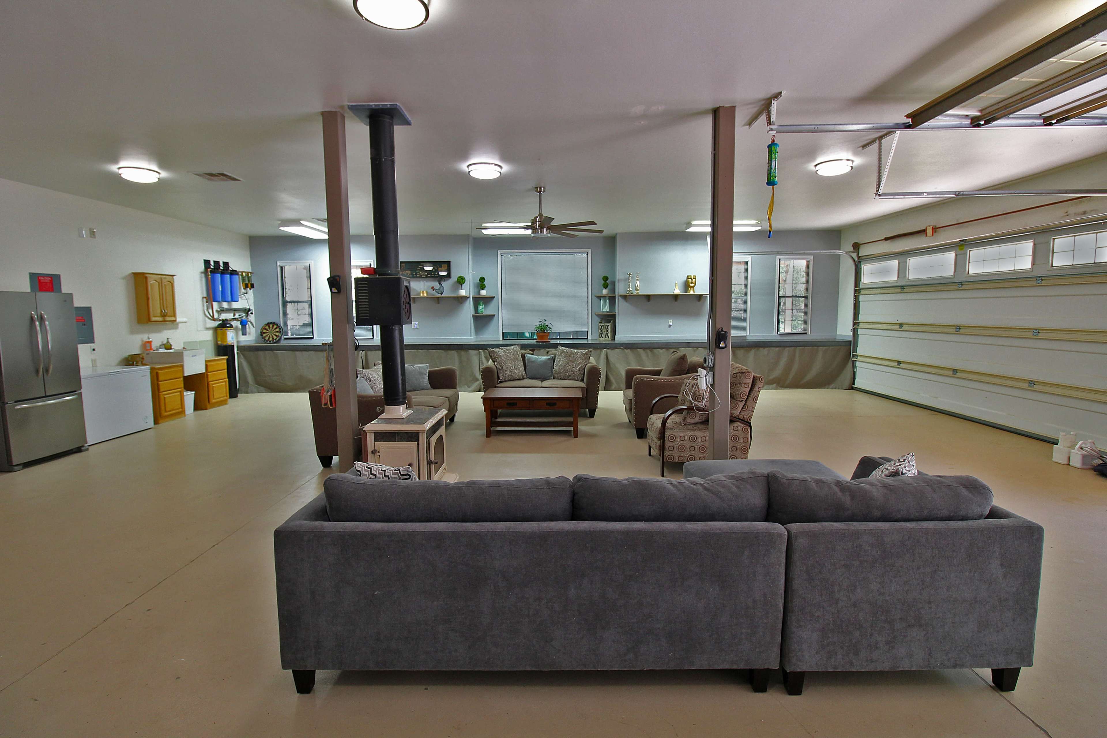A spacious garage converted into a living area, featuring a sectional couch, coffee table, and a kitchen area with cabinets and appliances.