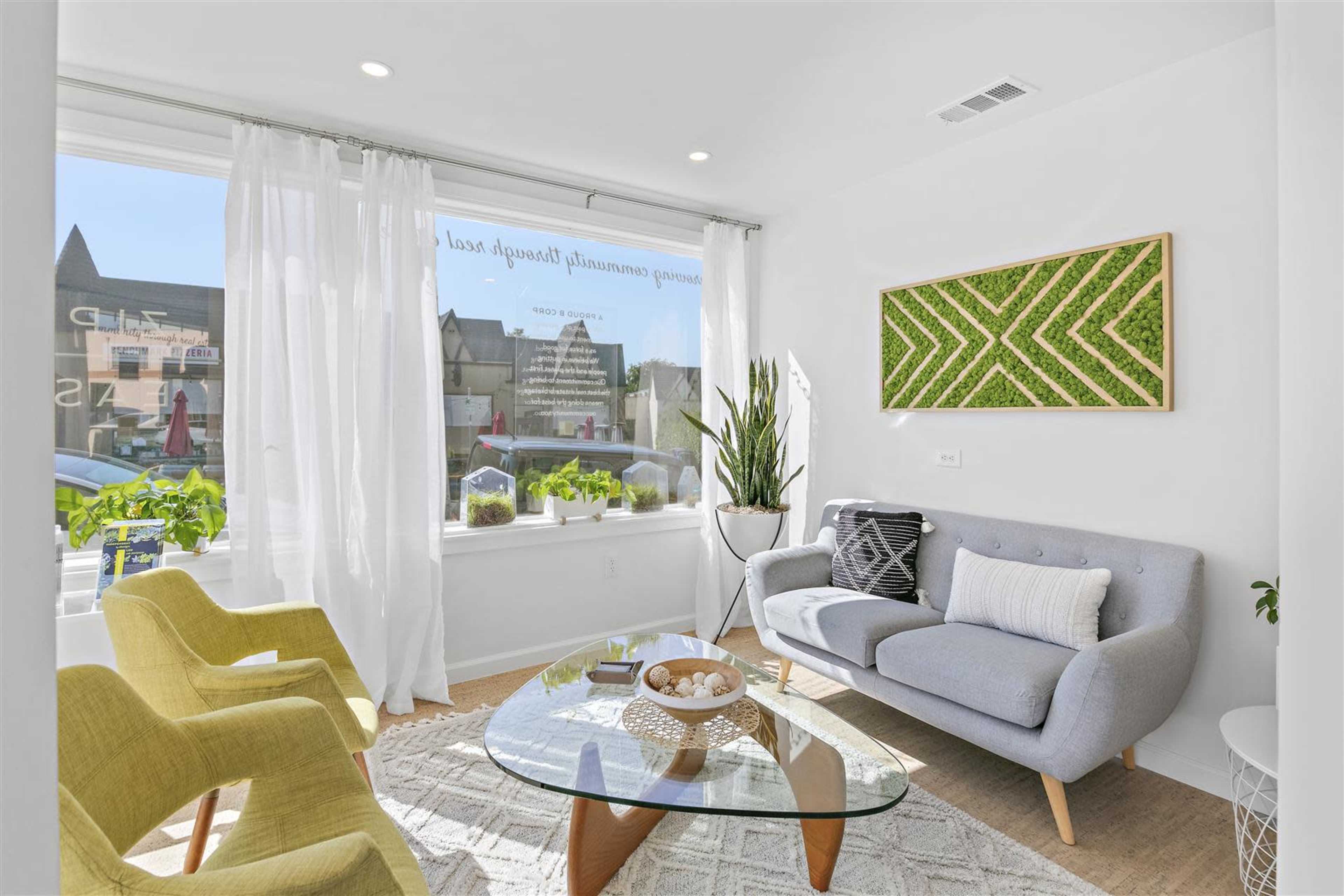A modern waiting room features a gray sofa, two yellow chairs, a glass coffee table, and a large window with plants and natural light.