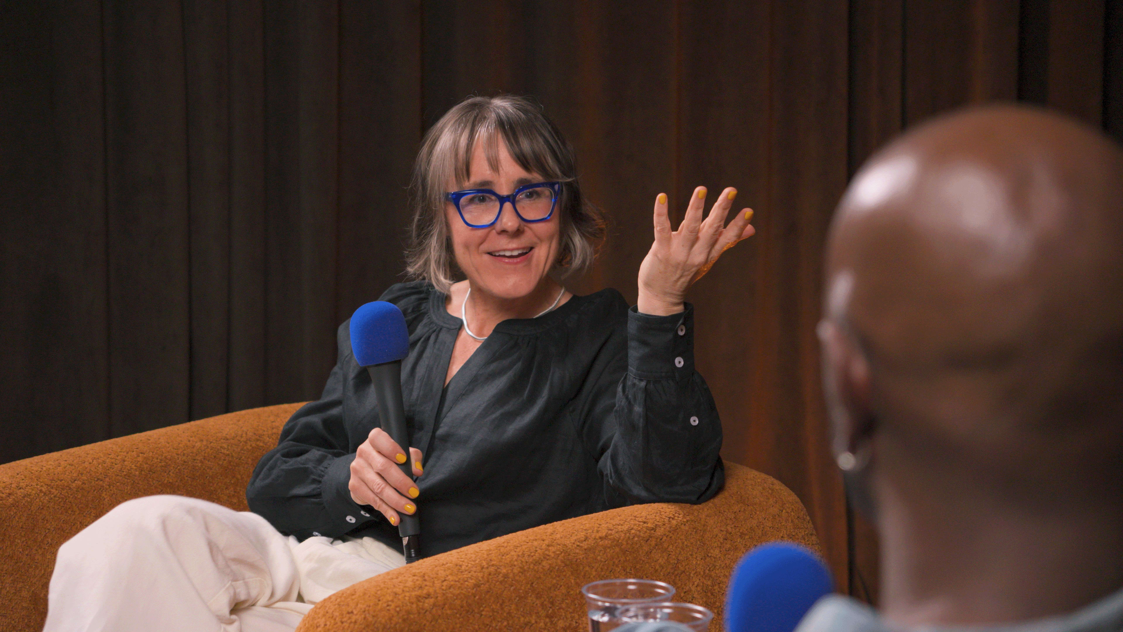 A woman with short hair and glasses is speaking into a microphone while seated on a brown couch during a conversation.