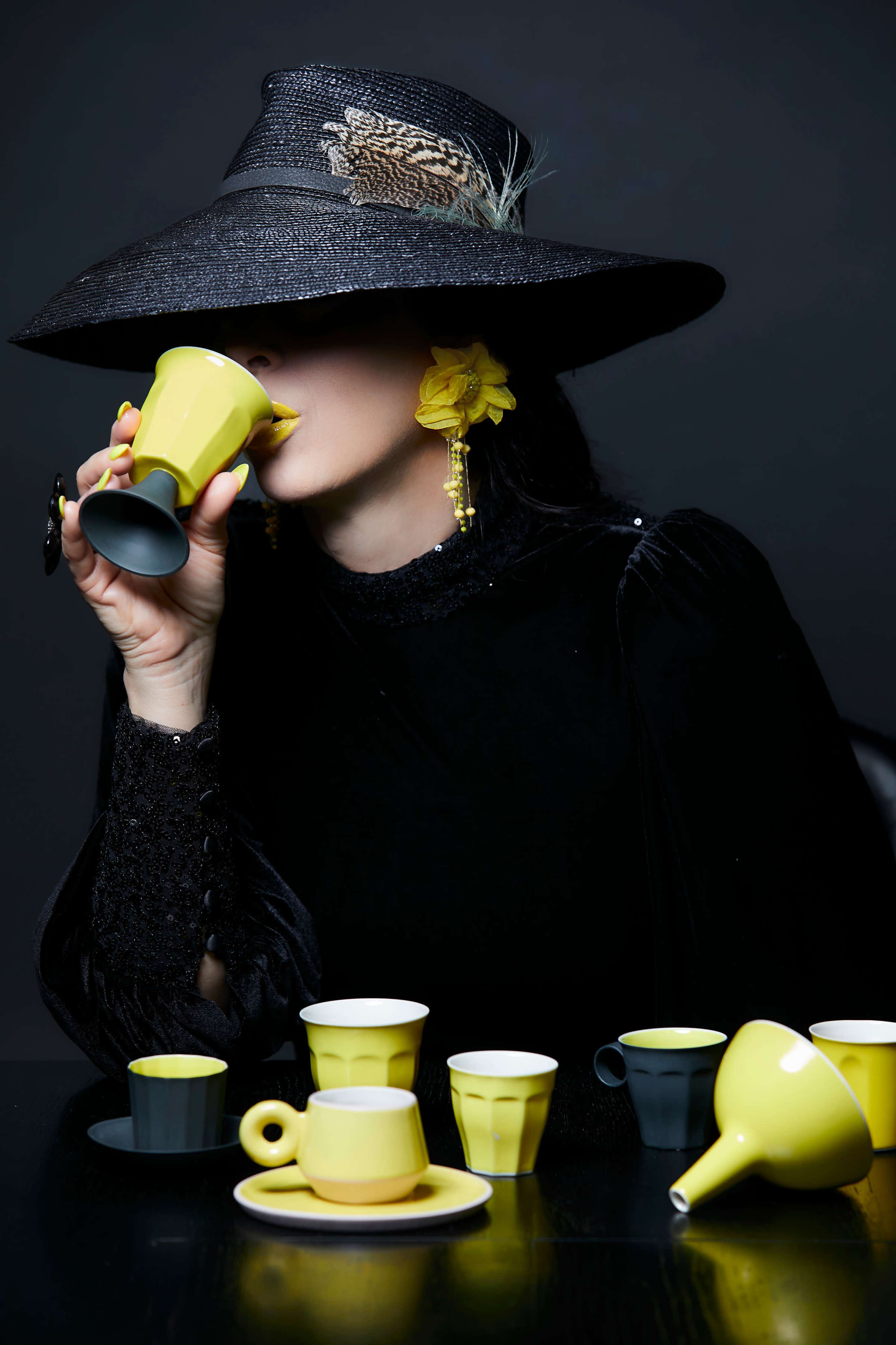 A woman wearing a large black hat sips from a yellow cup while surrounded by various yellow and gray ceramic cups on a dark table.