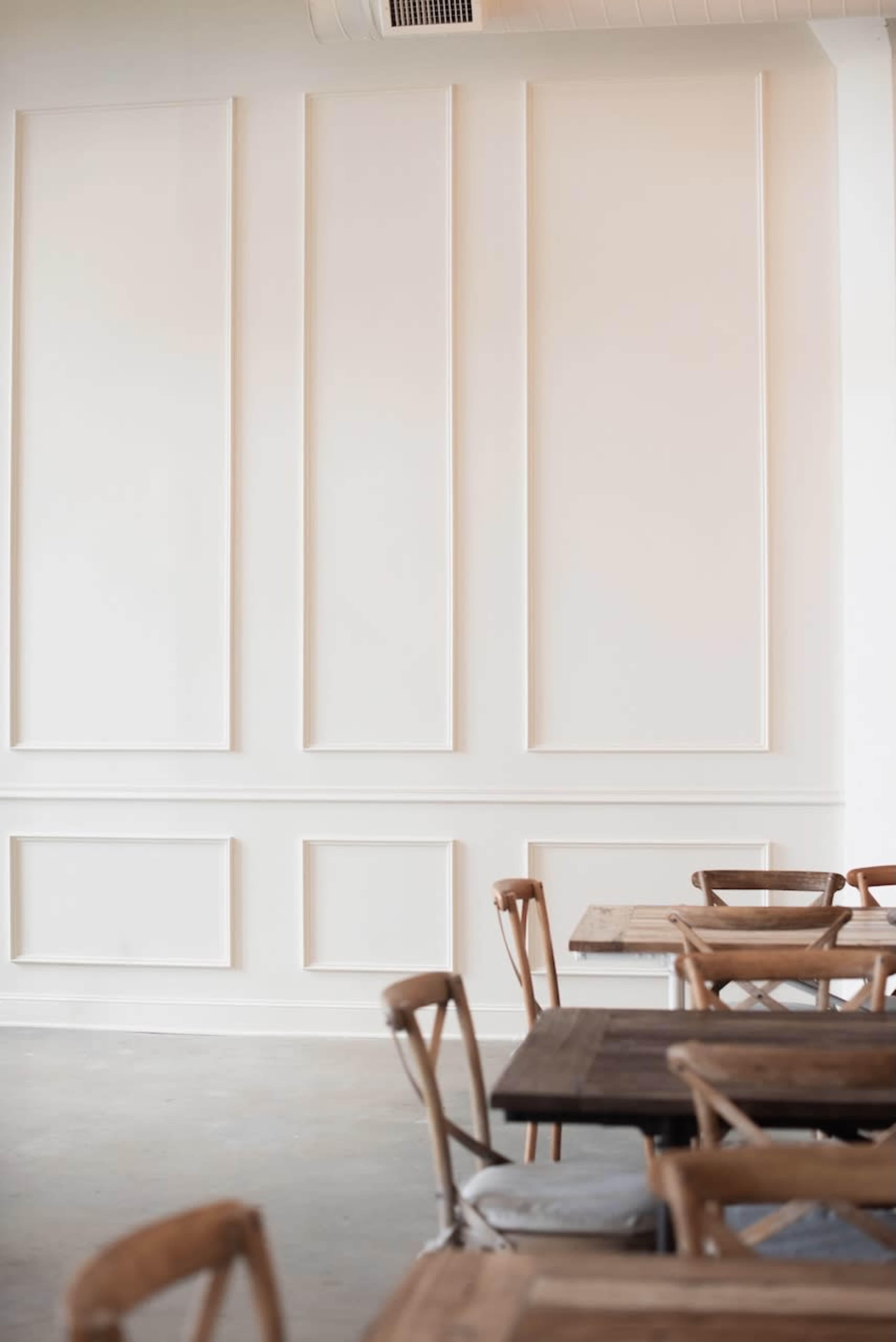 The image shows a minimalist interior of a cafe featuring white textured walls and wooden tables with chairs.