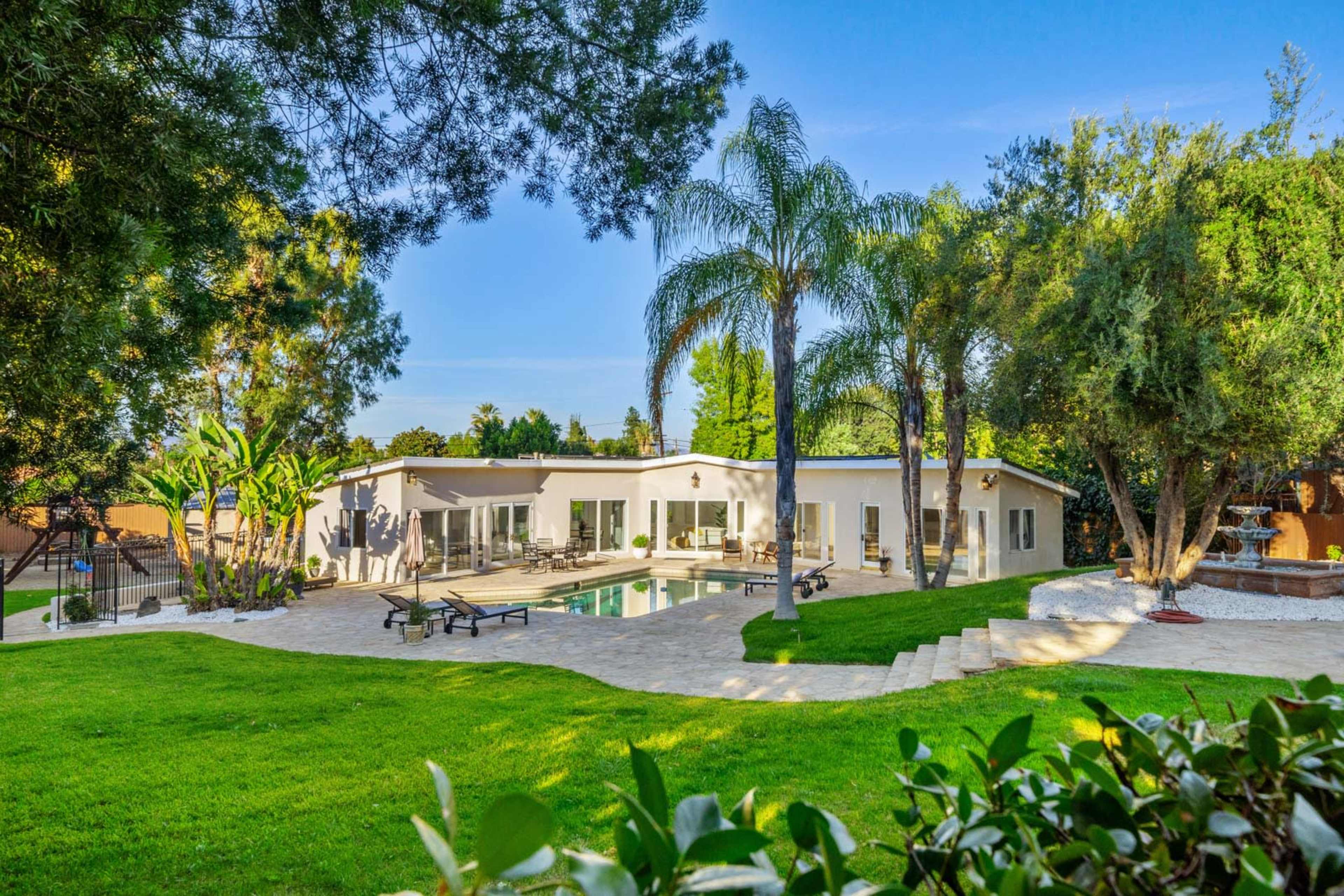 A modern house with large windows and a swimming pool surrounded by lush greenery and palm trees.