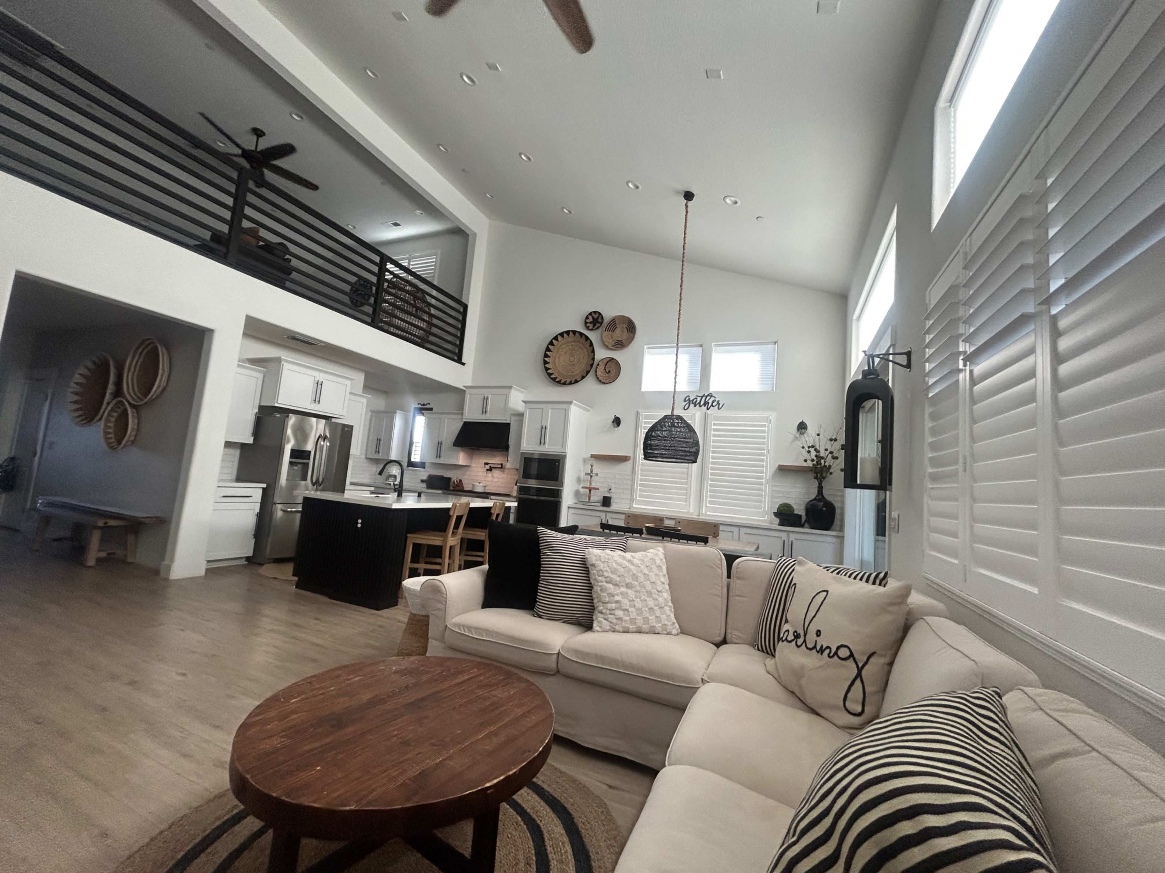 The image shows an open-concept living area with a light-colored sectional sofa, a round wooden coffee table, and a modern kitchen visible in the background.