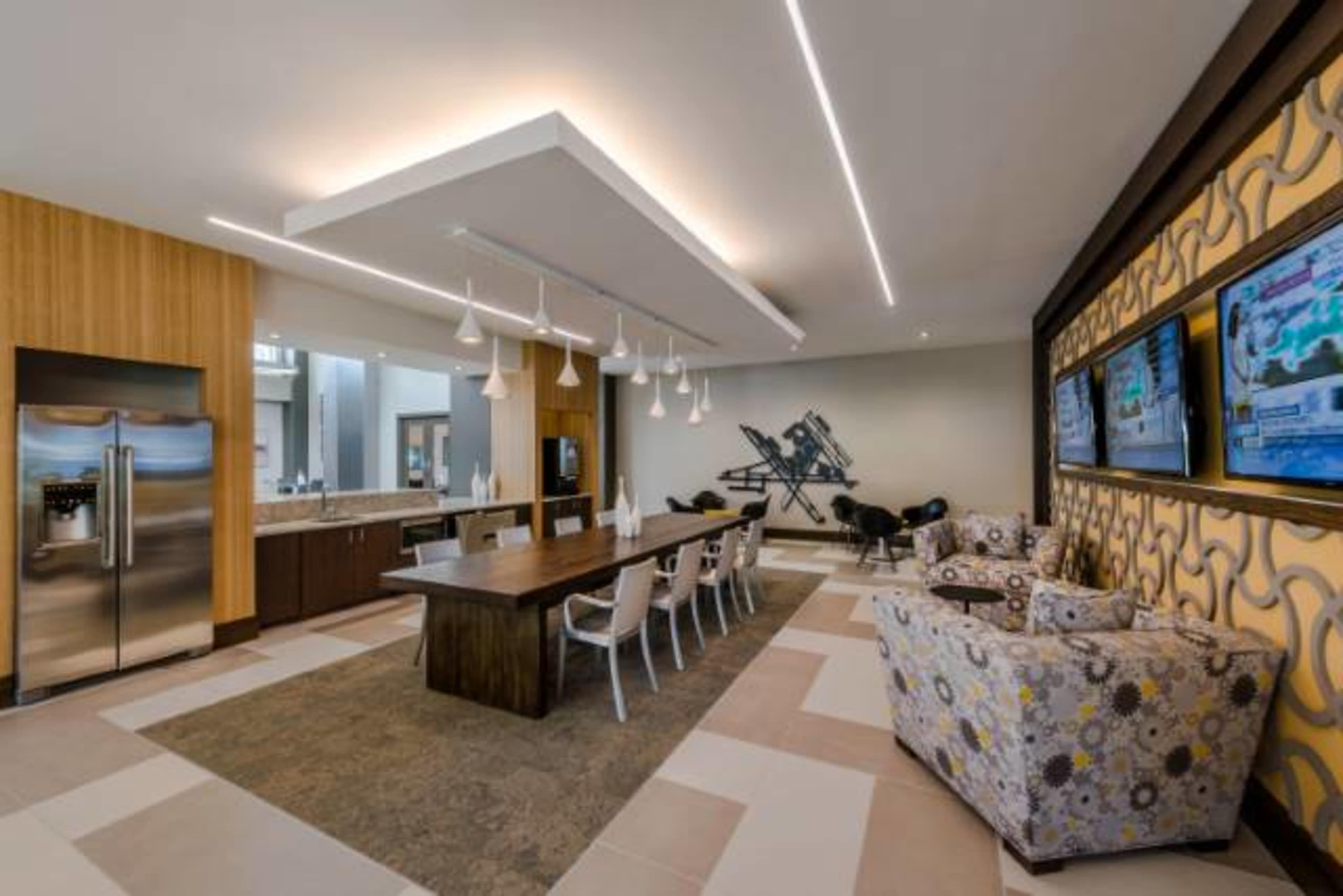 A modern dining area with a long wooden table, white chairs, and a kitchen space with stainless steel appliances, featuring television screens on the wall.