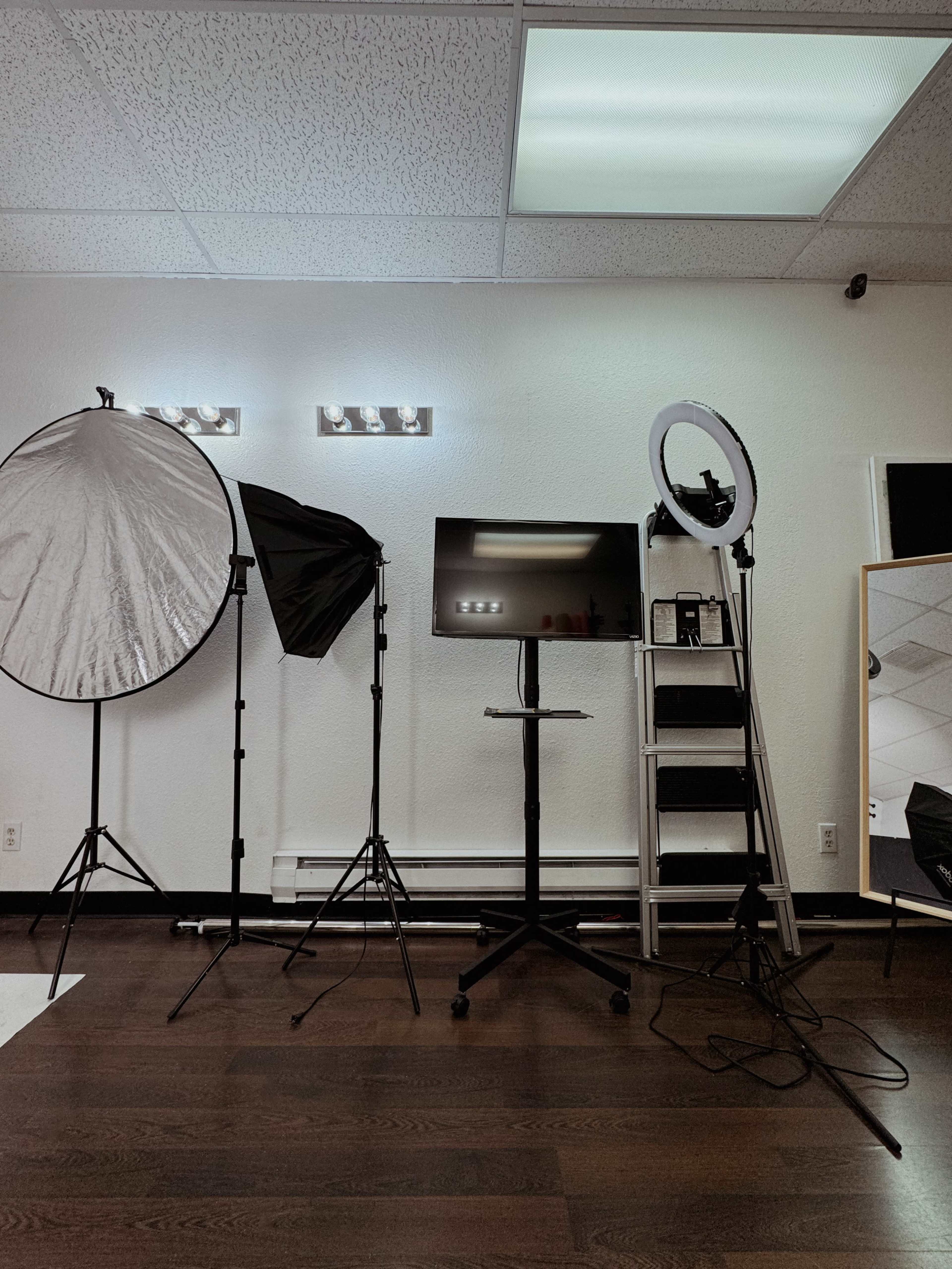 The image shows a photography setup featuring softboxes, a ring light, a monitor on a stand, and a mirror against a white wall.