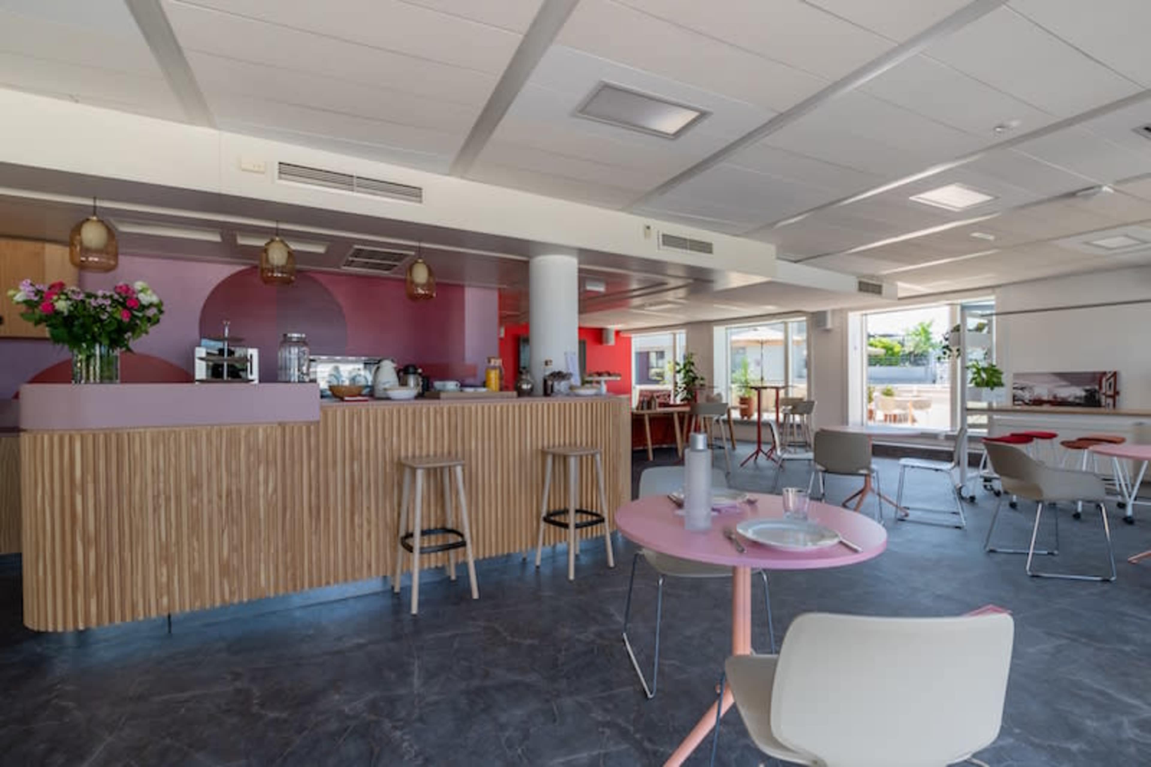 A modern café interior with a wooden counter, pink accents, and several tables and chairs arranged in a spacious layout.