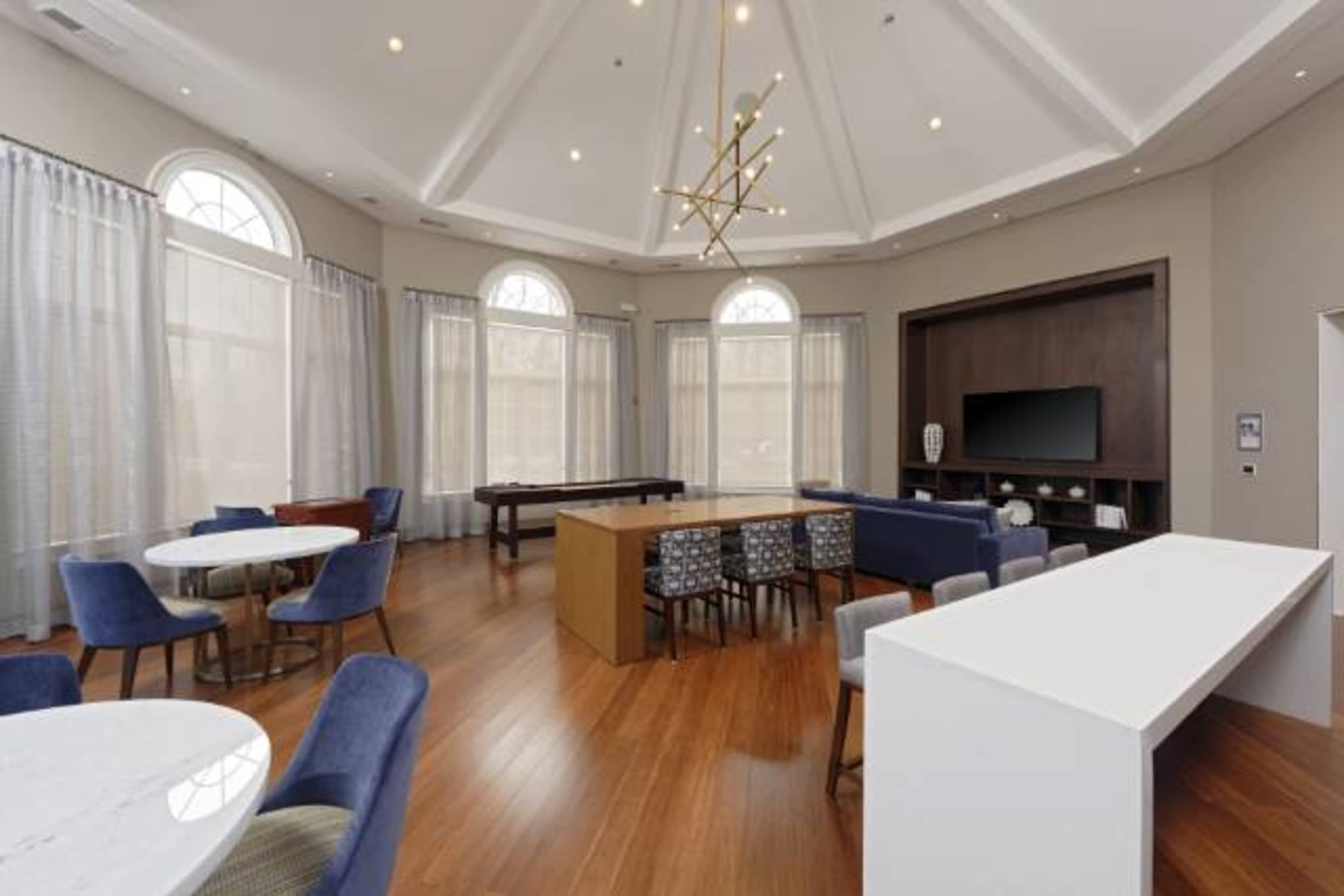 A spacious room with a high ceiling, featuring a seating area with multiple tables and chairs, large windows, and a media console with a television.