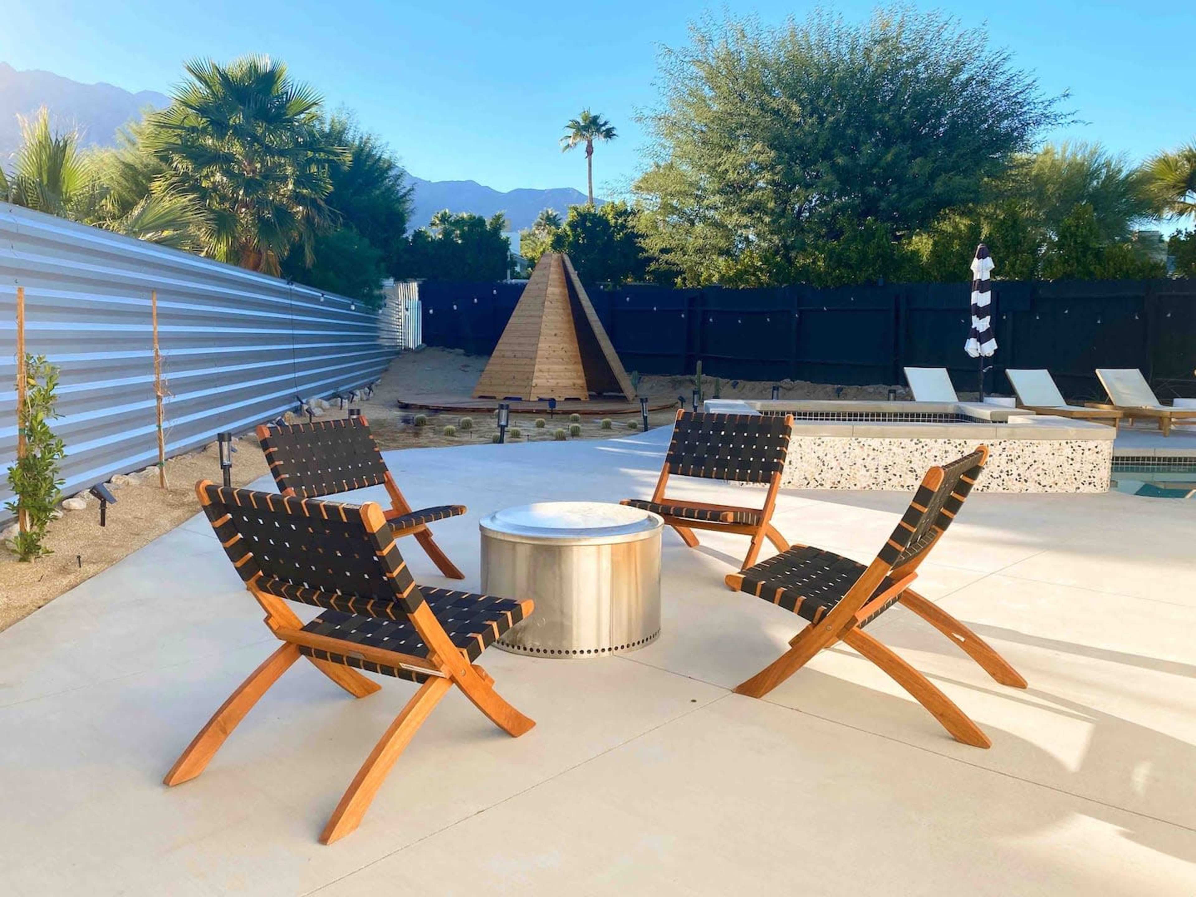 The scene features four wooden lounge chairs arranged around a circular fire pit beside a swimming pool, with palm trees and a pyramid-shaped structure in the background.