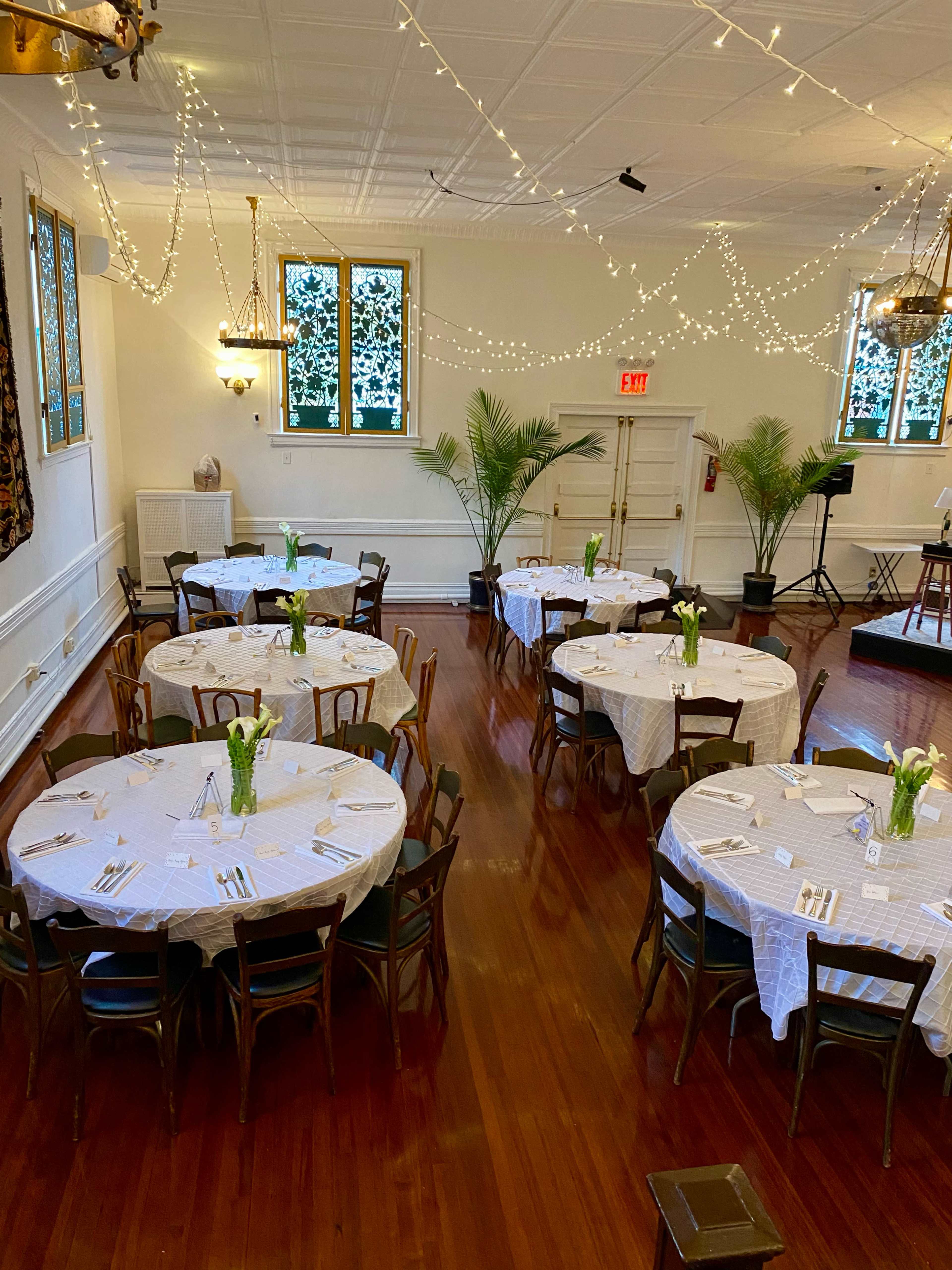 A banquet hall with several round tables set with white tablecloths and centerpieces, illuminated by soft string lights and flanked by potted plants.