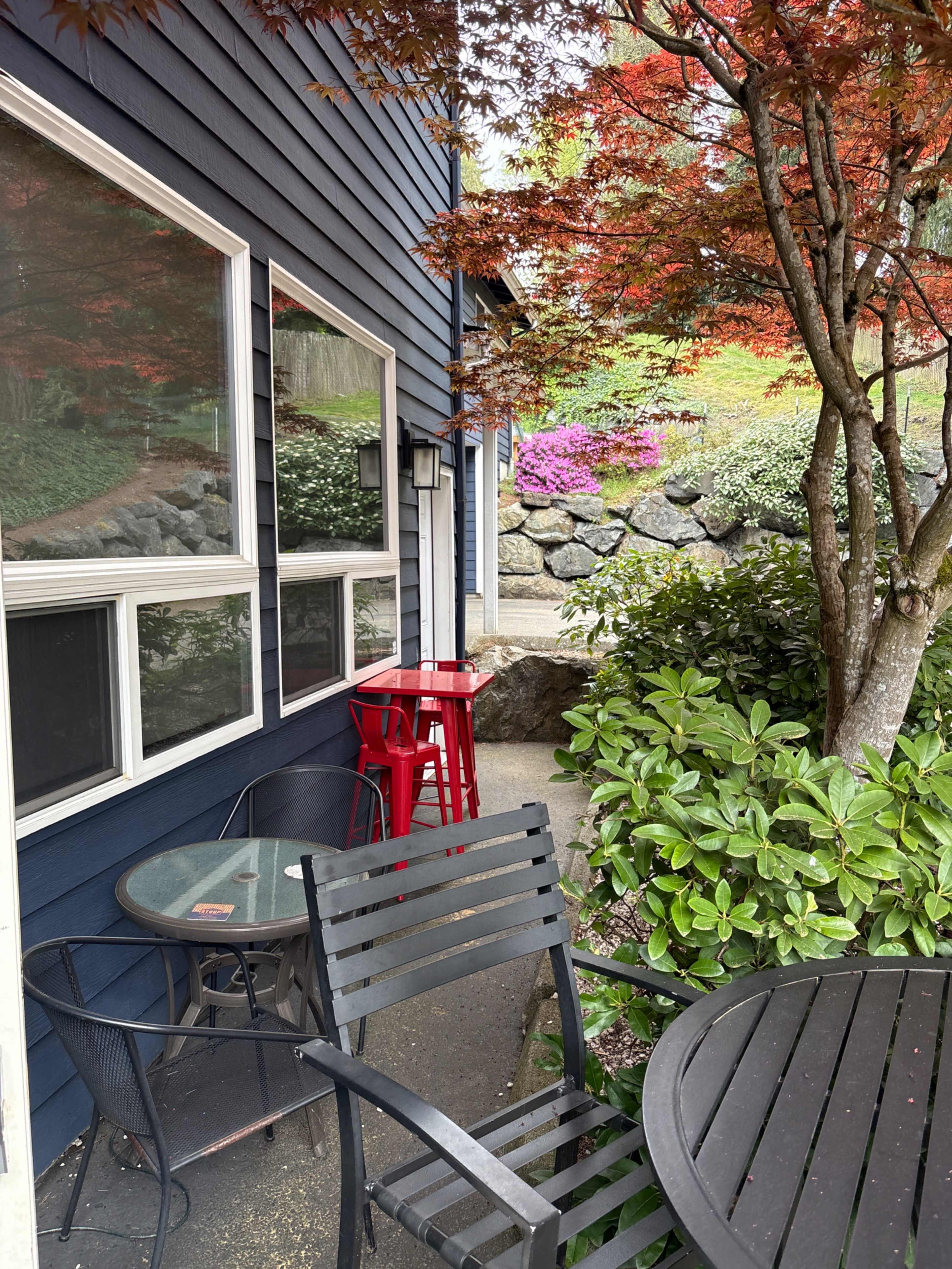 The image shows a patio area with a black table and chairs, a red stool, and a backdrop of green foliage and colorful flowers beside a blue house.