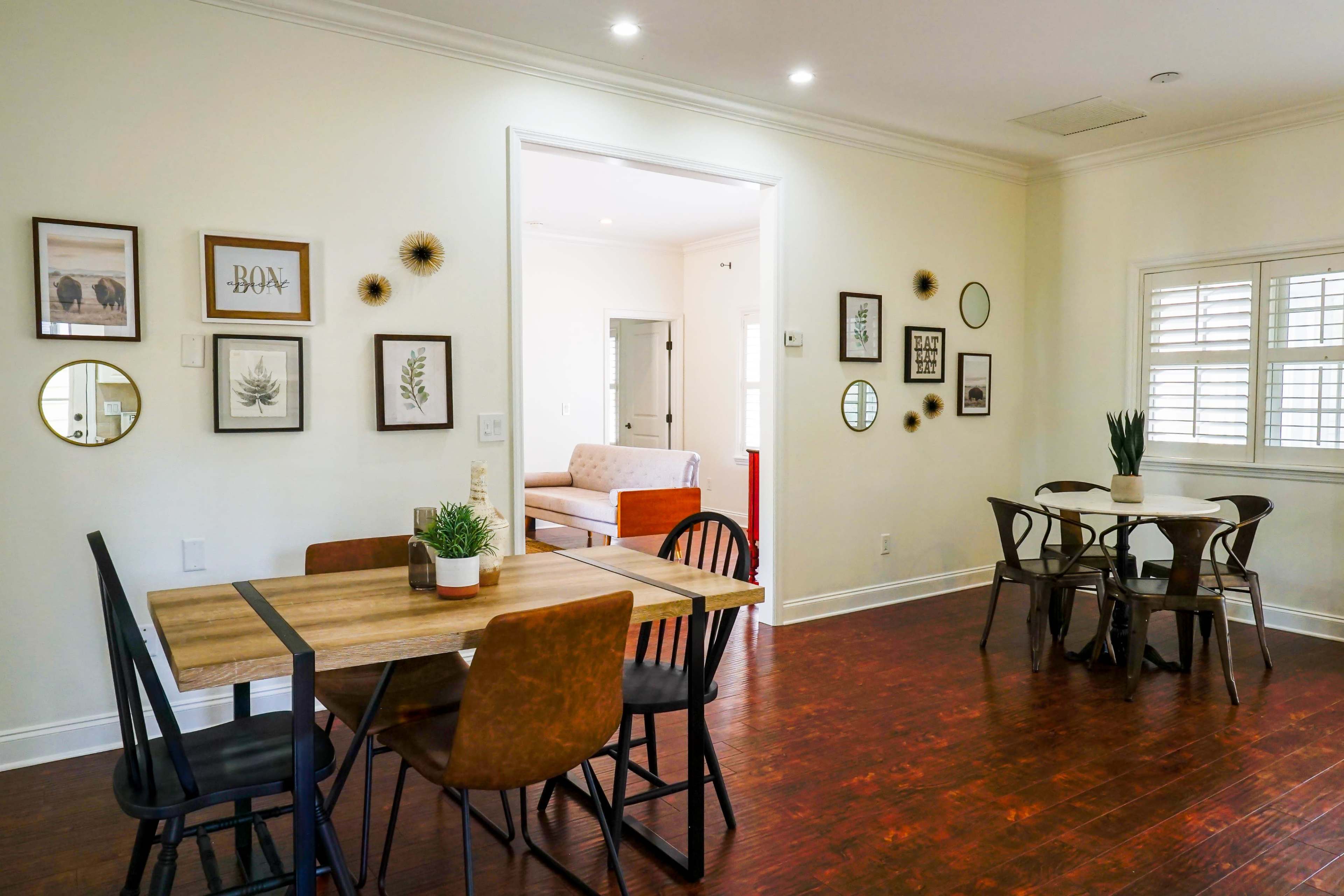 A dining area features a wooden table with chairs, surrounded by framed wall art and mirrors, leading to a living room with a sofa in the background.