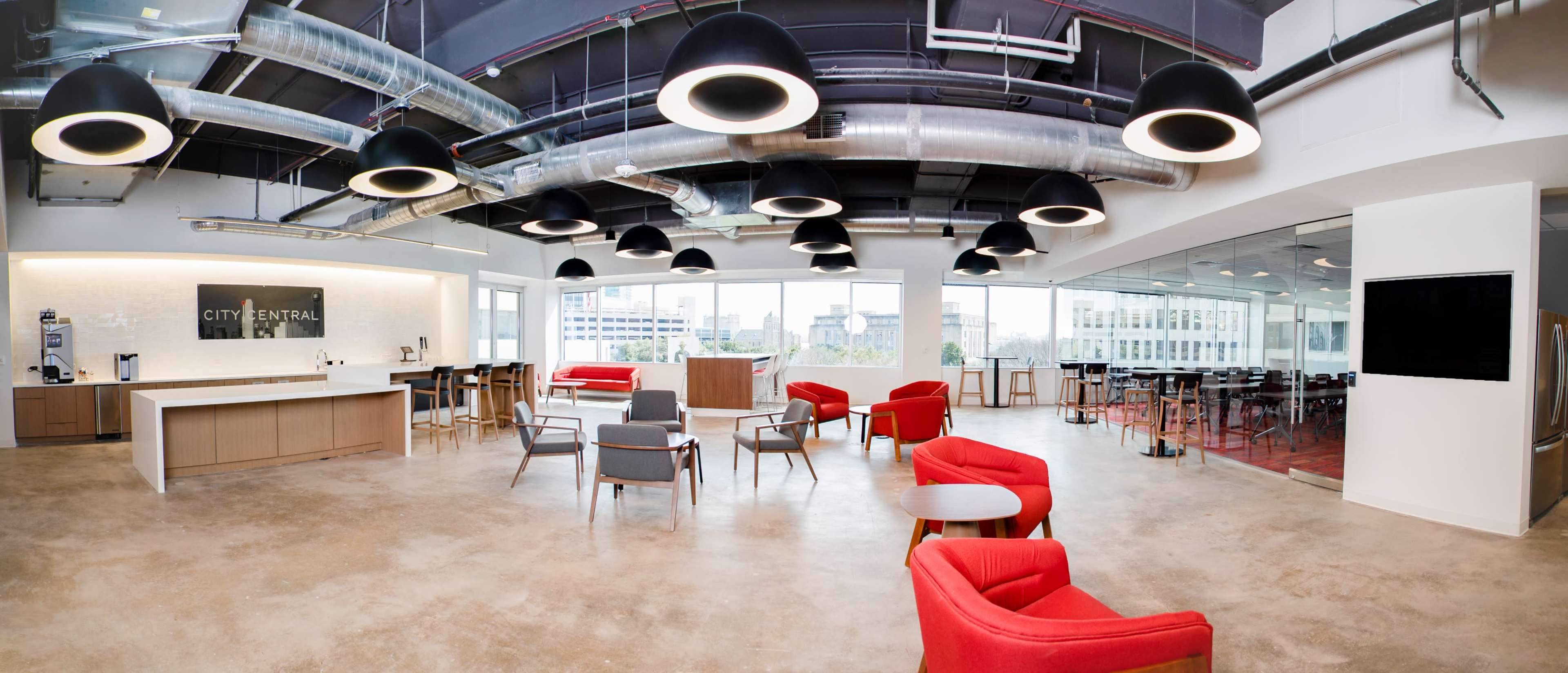 A modern office space with red lounge chairs, a kitchenette area, and large windows letting in natural light.