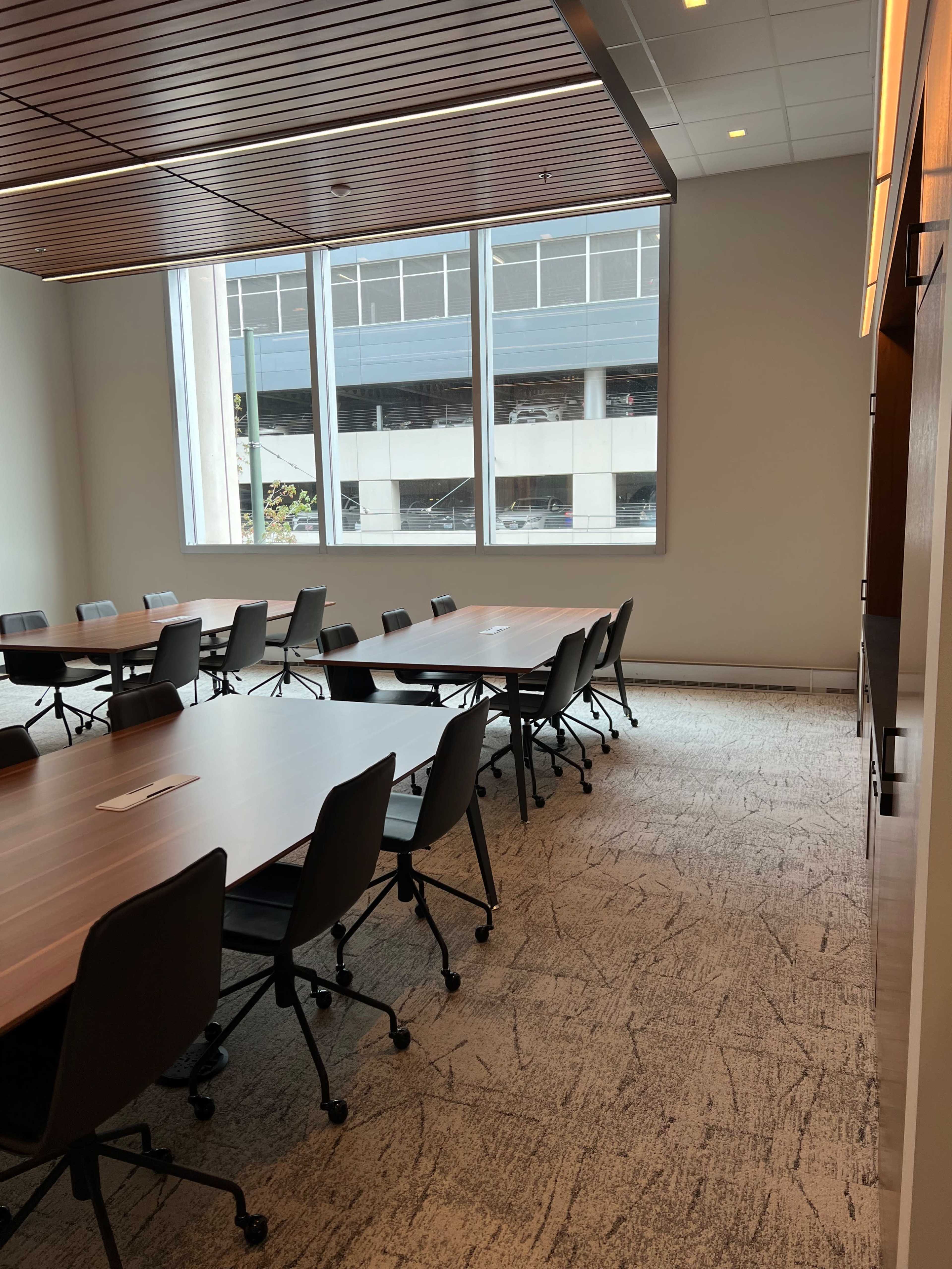The image shows a modern conference room with several large tables arranged for meetings, surrounded by chairs and large windows allowing natural light to enter.