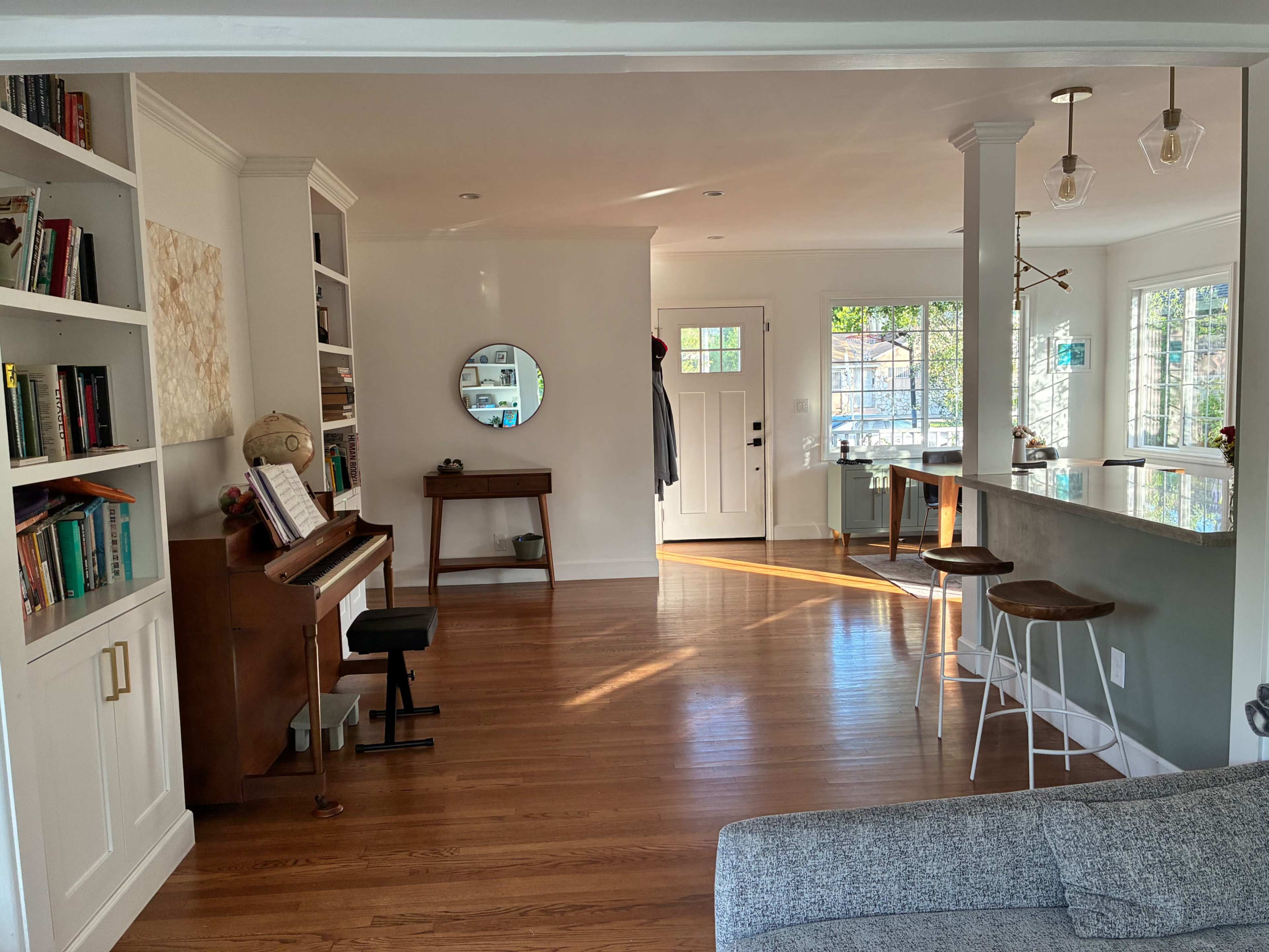 A living area features a piano beside a bookshelf, a dining table with chairs in view, and large windows that allow natural light to fill the space.