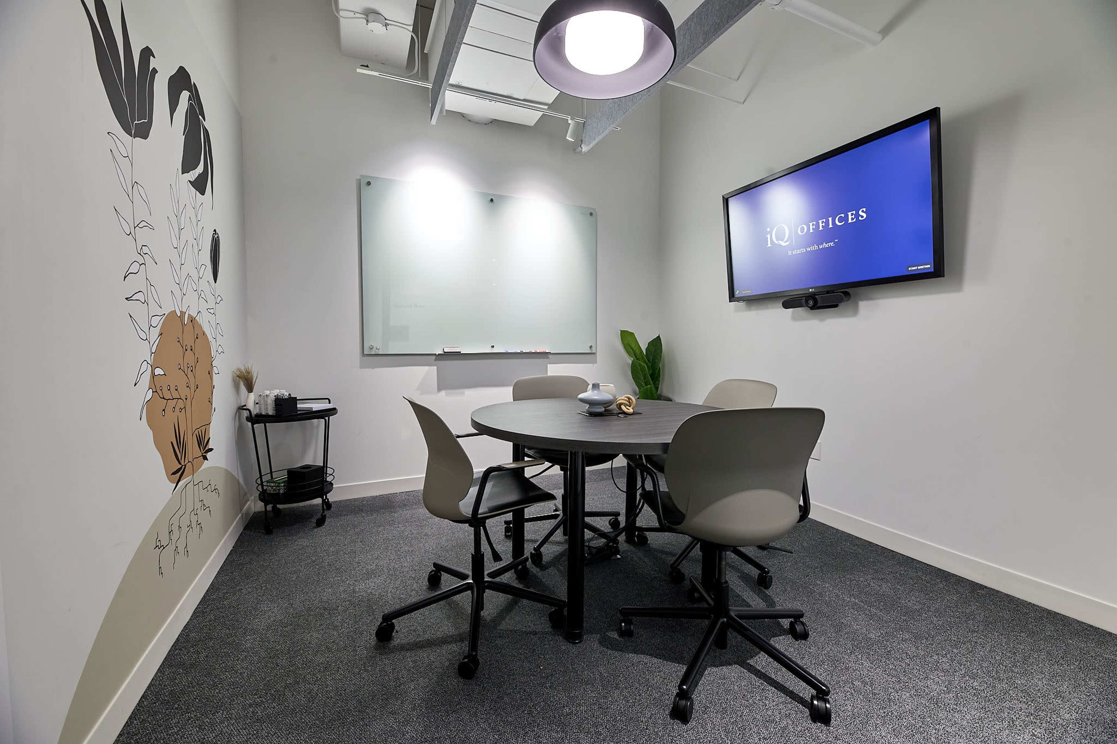 A small meeting room features a round table with four chairs, a wall-mounted screen, a whiteboard, and a decorative mural.