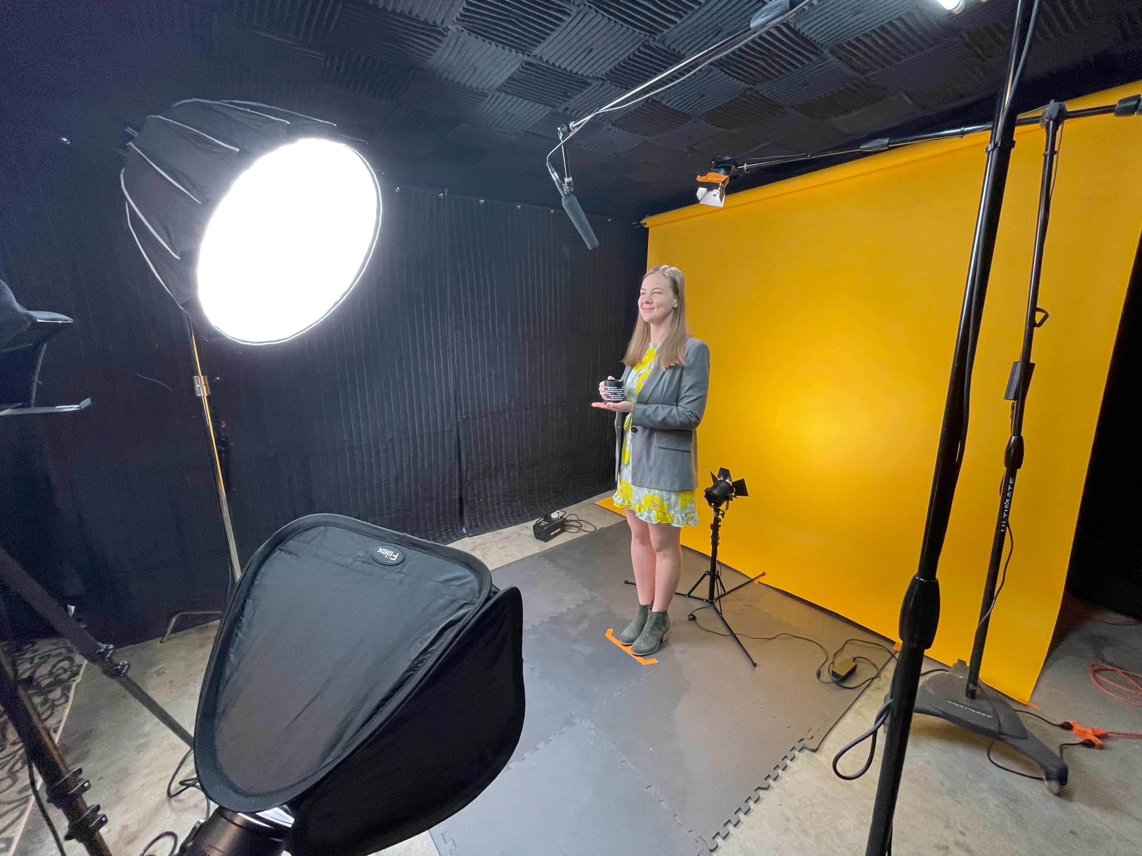 A woman stands in front of a bright yellow backdrop in a photo studio, holding a camera and illuminated by soft lighting.