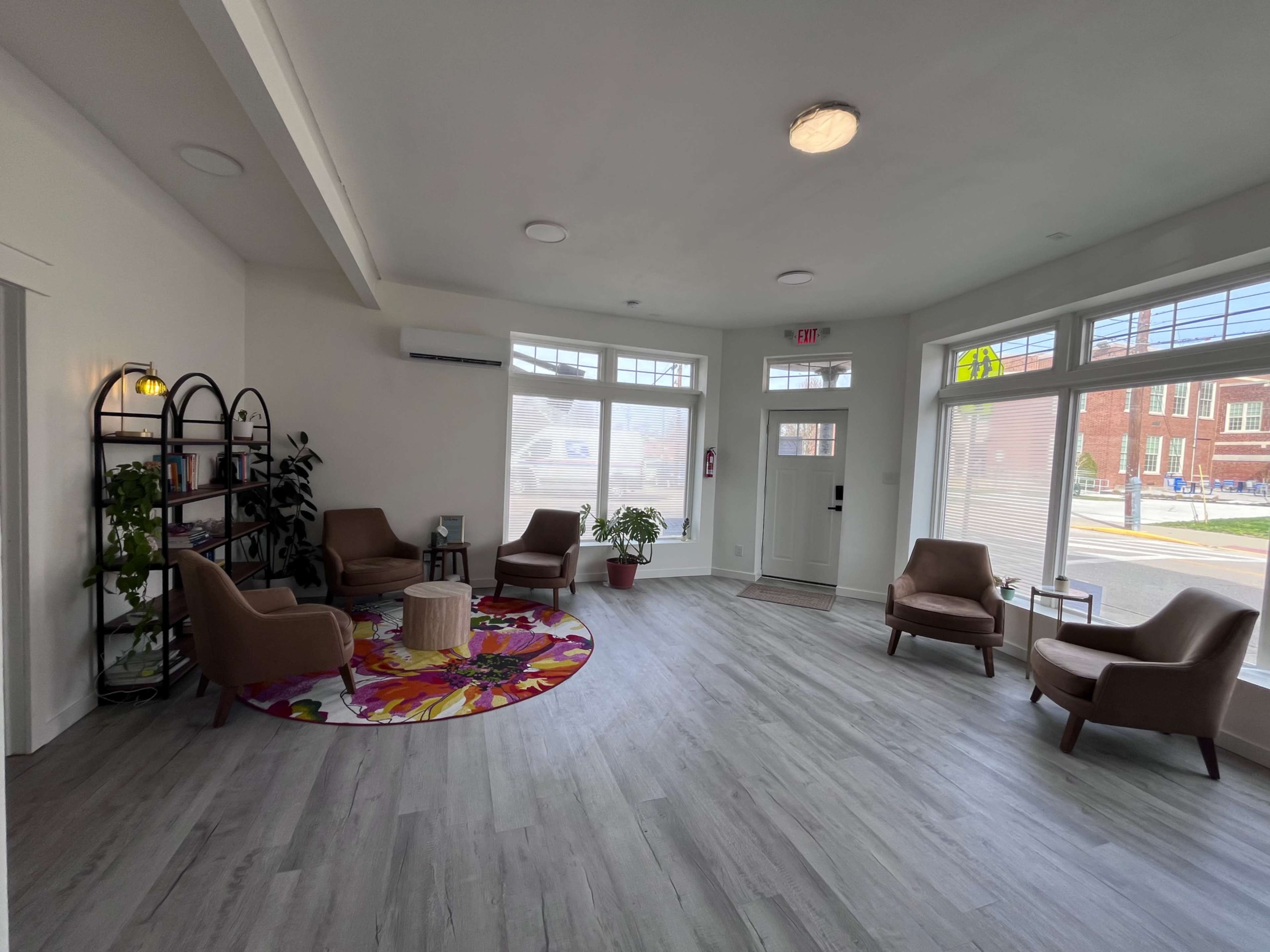 A bright, modern waiting area features four upholstered chairs arranged around a circular table, with a colorful rug and a bookshelf filled with plants.
