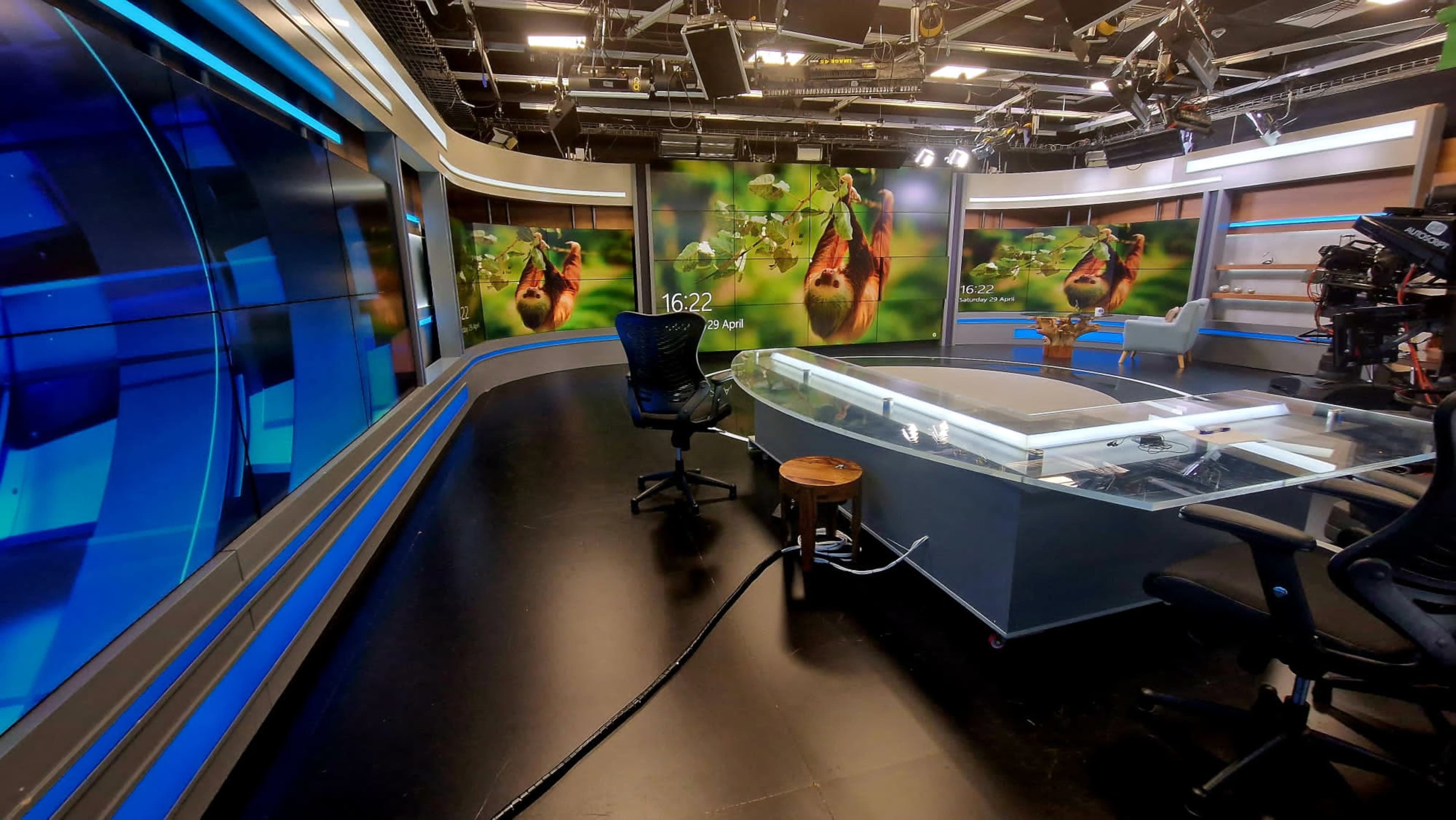The image shows a television studio with a glass news desk, black chairs, and large digital screens displaying a nature scene in the background.