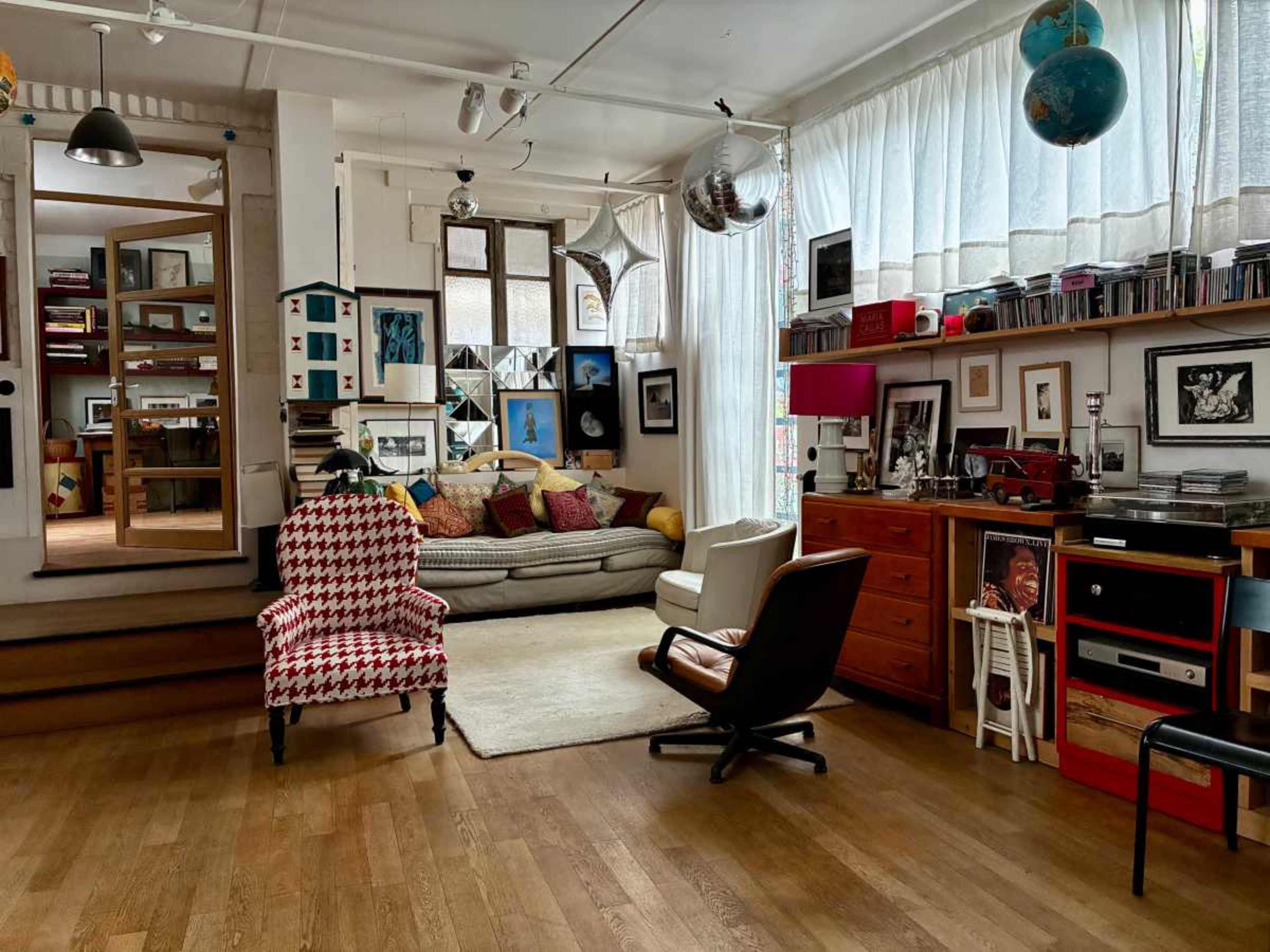 The image shows a cozy living space featuring a mix of furniture, bookshelves filled with books and art, and large windows allowing natural light to enter.