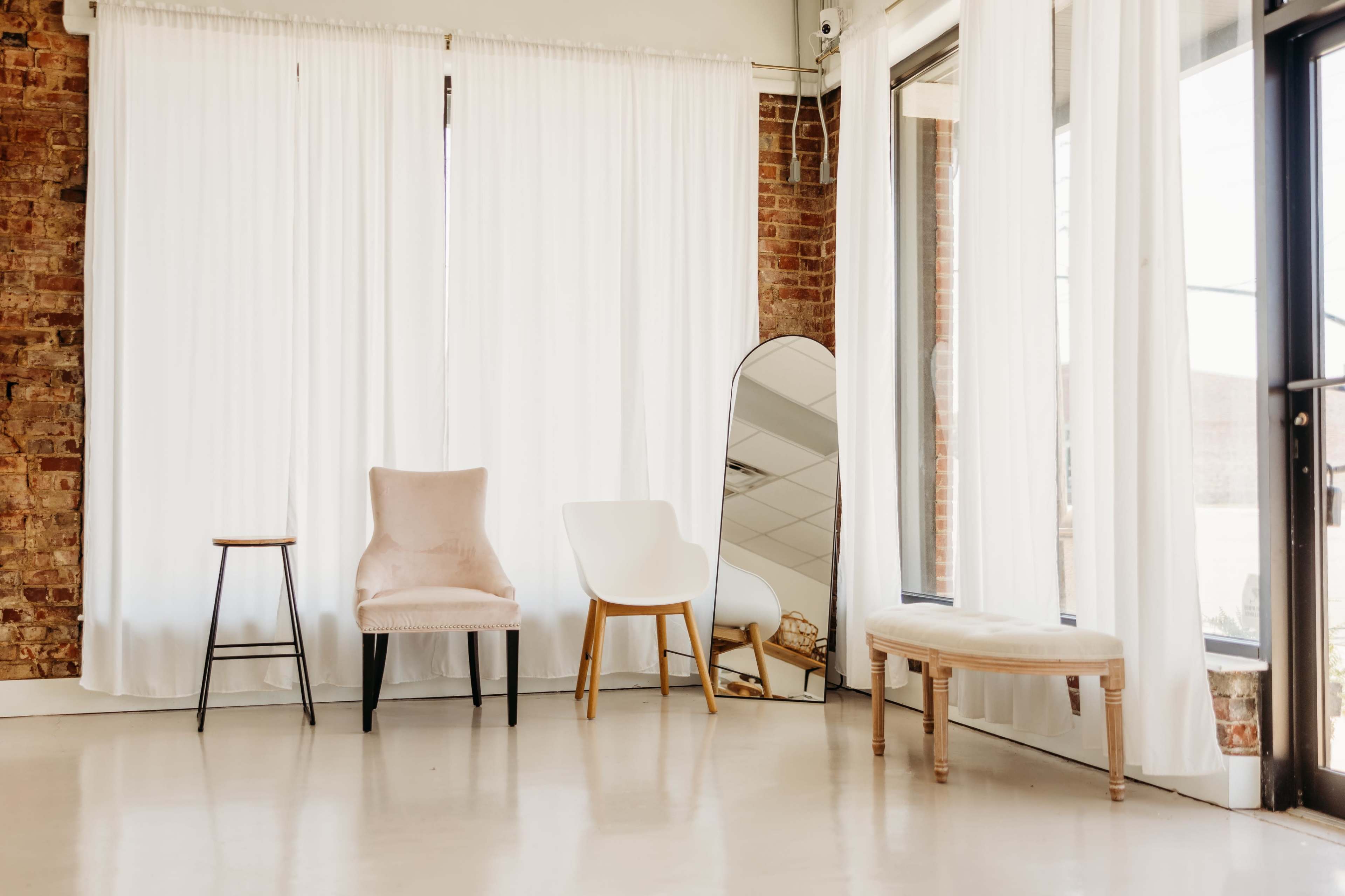 The scene shows a well-lit room with three chairs of different styles, a large mirror, and white curtains against a brick wall.