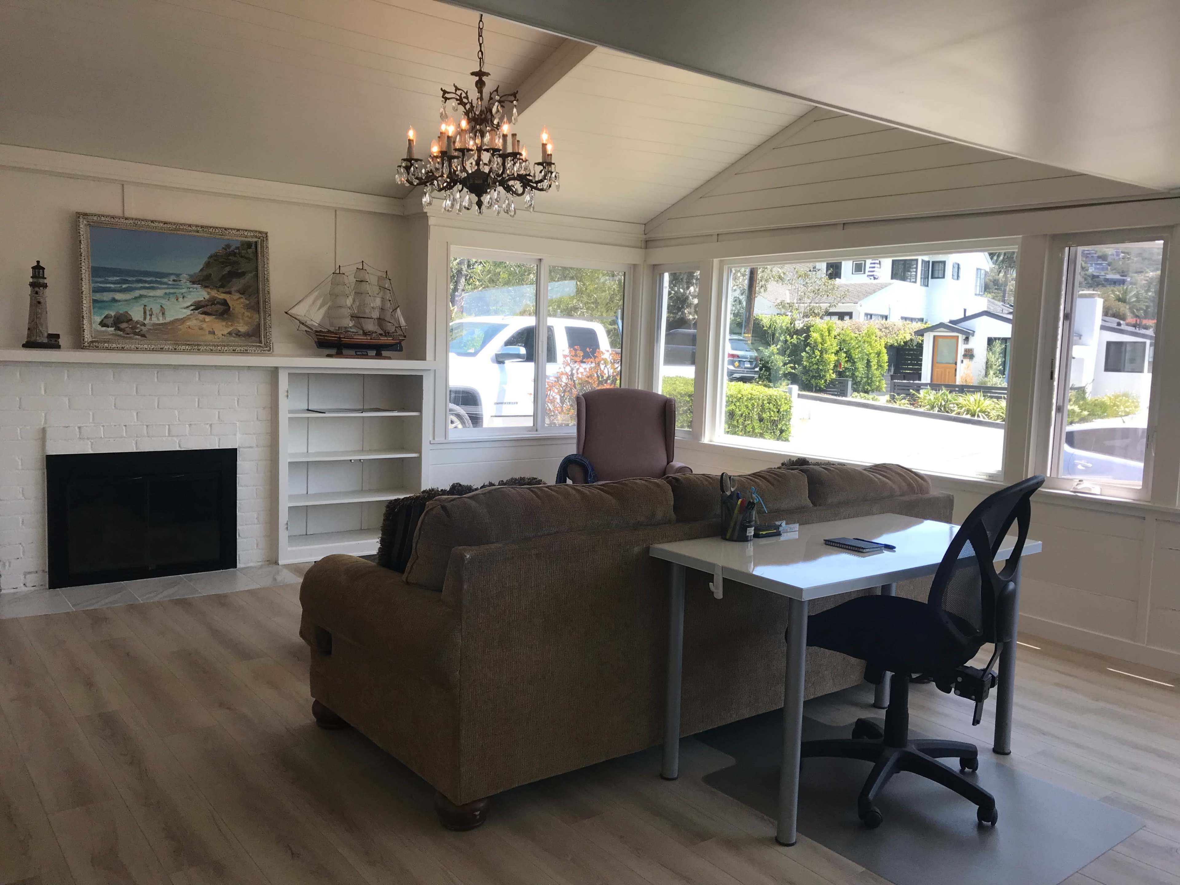 A bright, open living room with a chandelier, a couch facing a fireplace, and a desk with a chair near large windows.