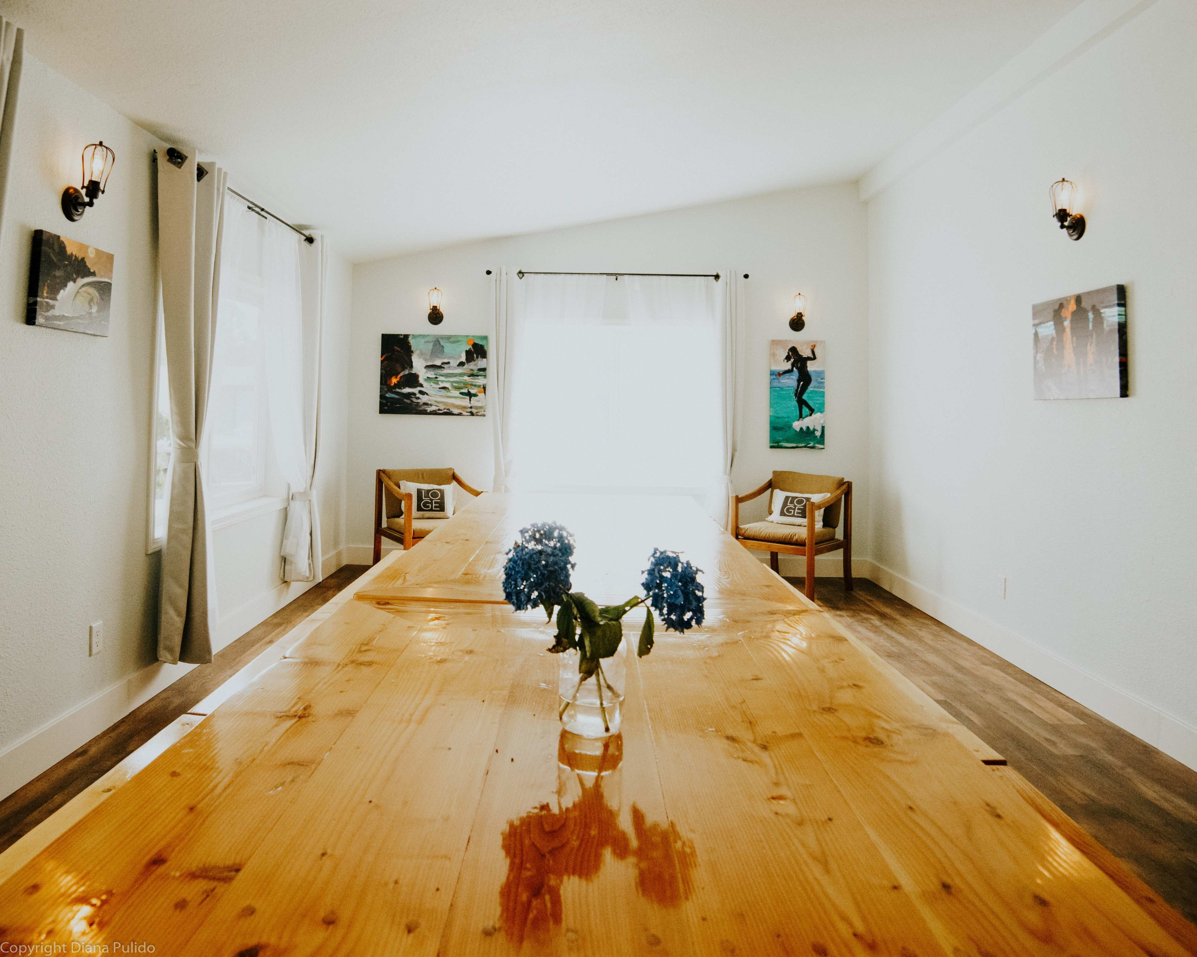 A spacious dining room features a large wooden table with a vase of blue flowers, flanked by two chairs and lit by wall sconces.