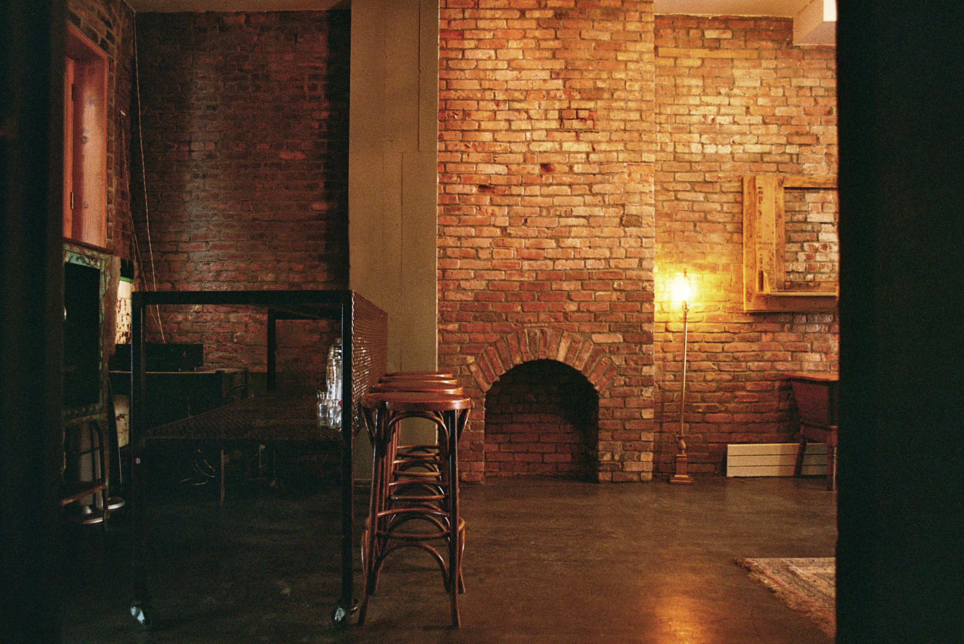The image shows a room with exposed brick walls, a metal bar with stools, and a warm lamp in the corner.