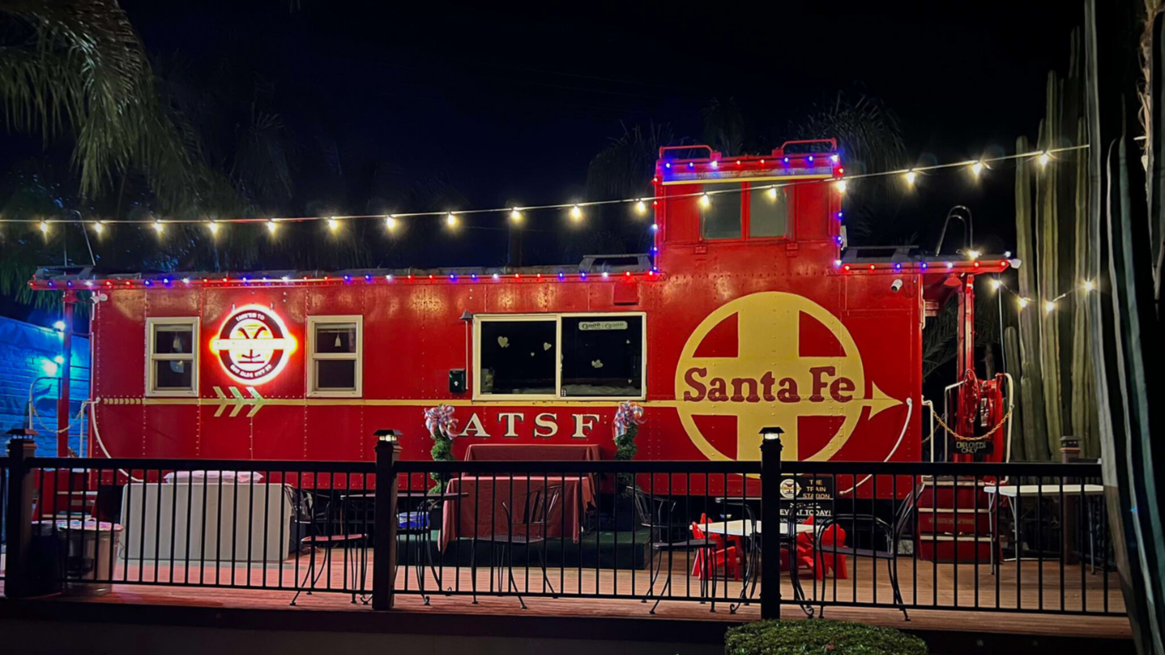 A brightly lit red Santa Fe train caboose is displayed in a nighttime setting, surrounded by patio furniture and decorative string lights.