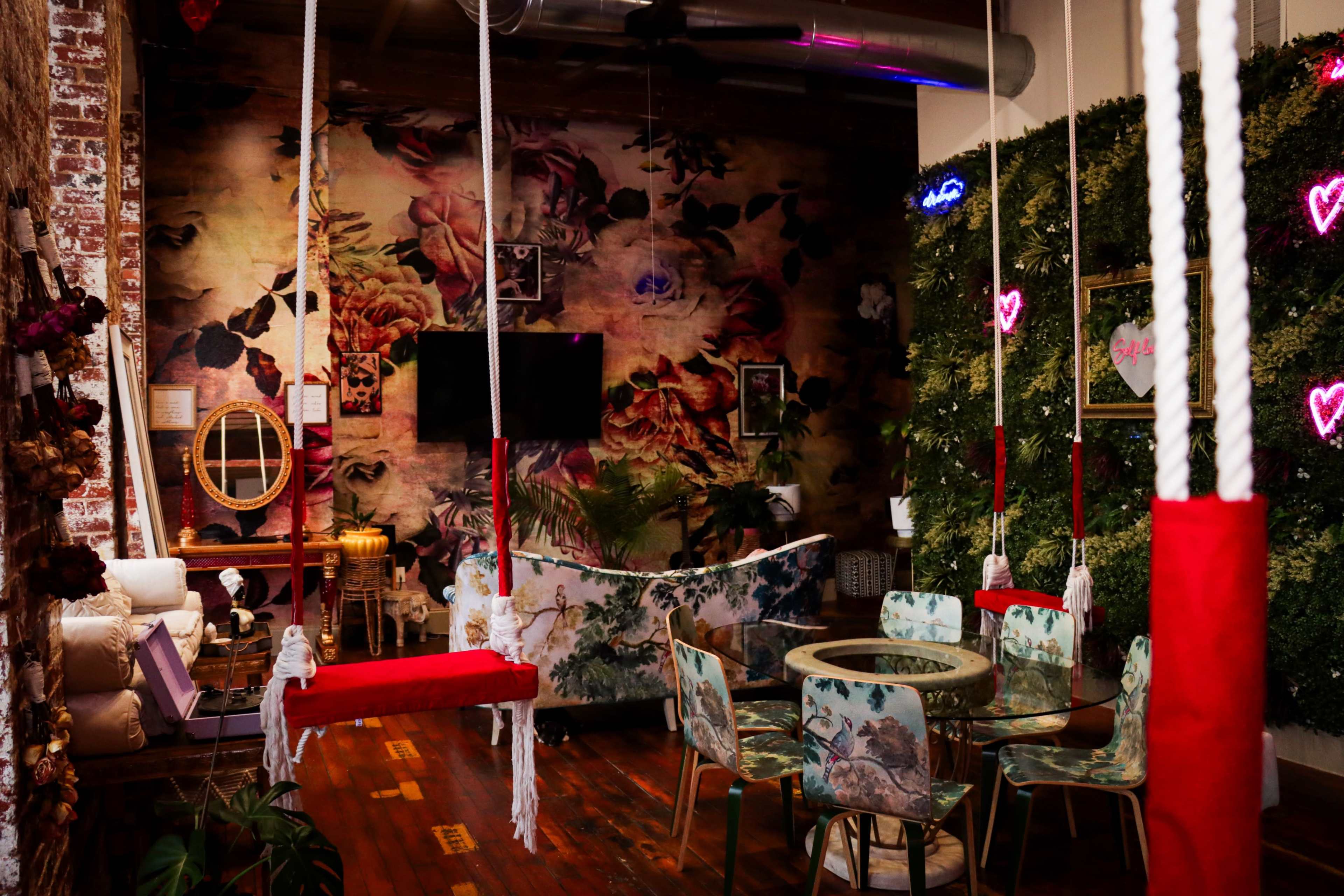 The scene features a stylish cafe with floral wallpaper, swinging seats, and a circular table surrounded by seating upholstered in a tropical pattern.
