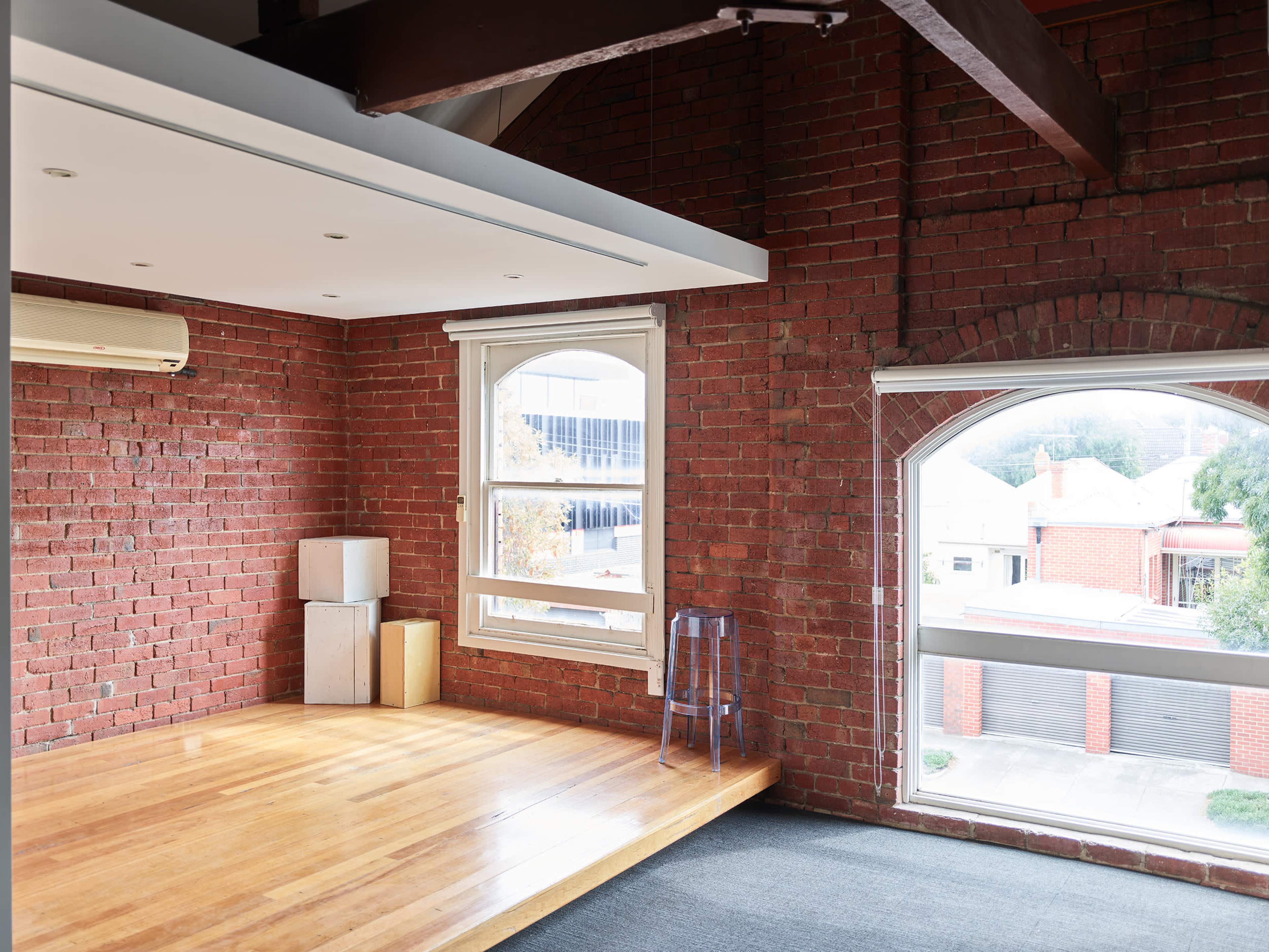 A spacious room features exposed brick walls, wooden flooring, and large windows that provide ample natural light.