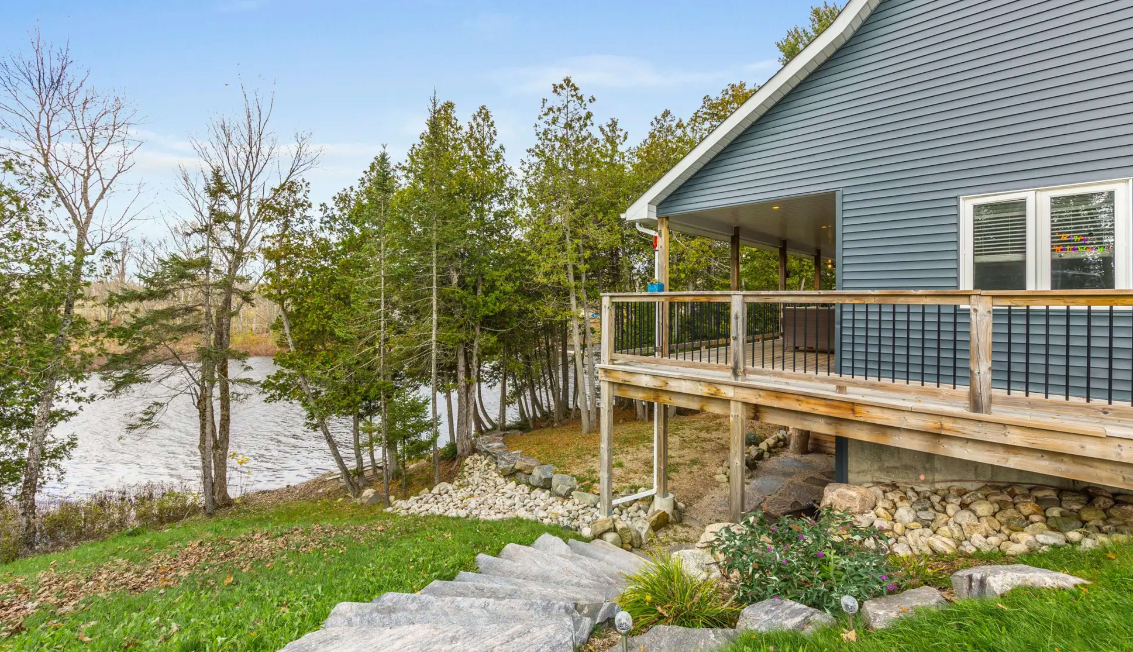 A blue house with a wooden deck overlooks a body of water, surrounded by trees and landscaped with stone steps and greenery.