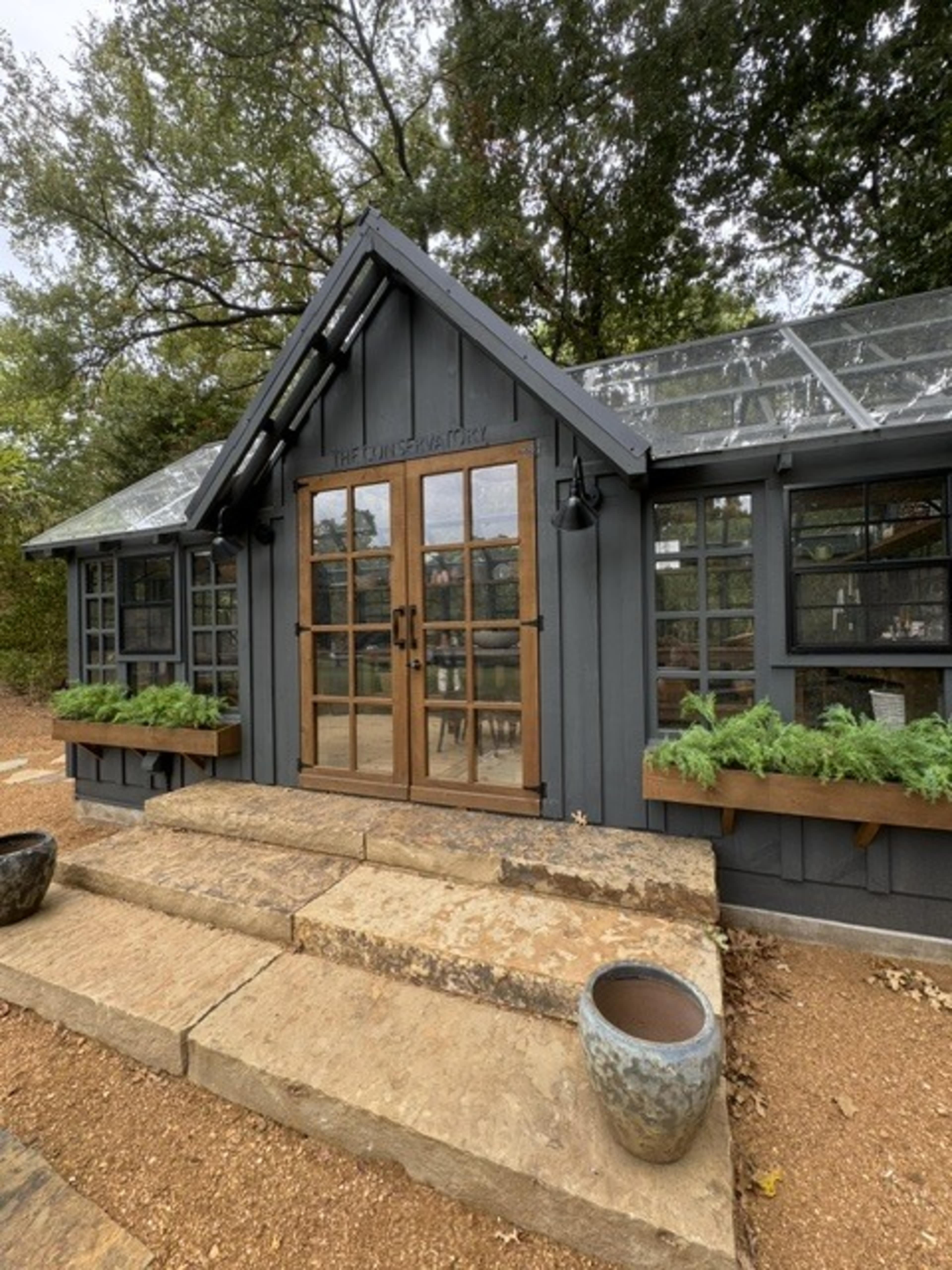 The image shows a modern gray garden conservatory with large glass windows and wooden doors, flanked by planters filled with greenery, set against a backdrop of trees.