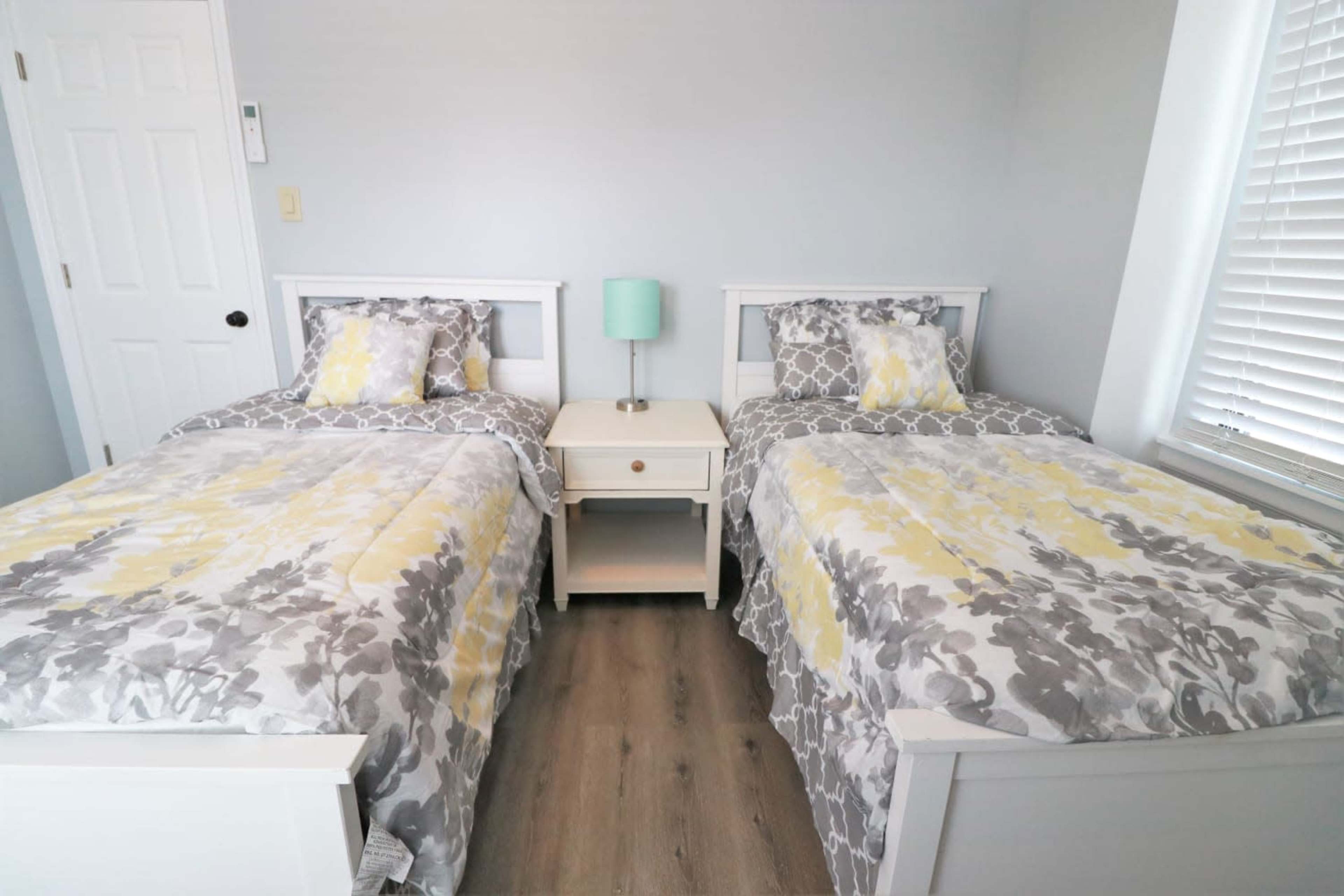 A bright bedroom featuring two twin beds with gray and yellow bedding, separated by a small nightstand with a teal lamp.
