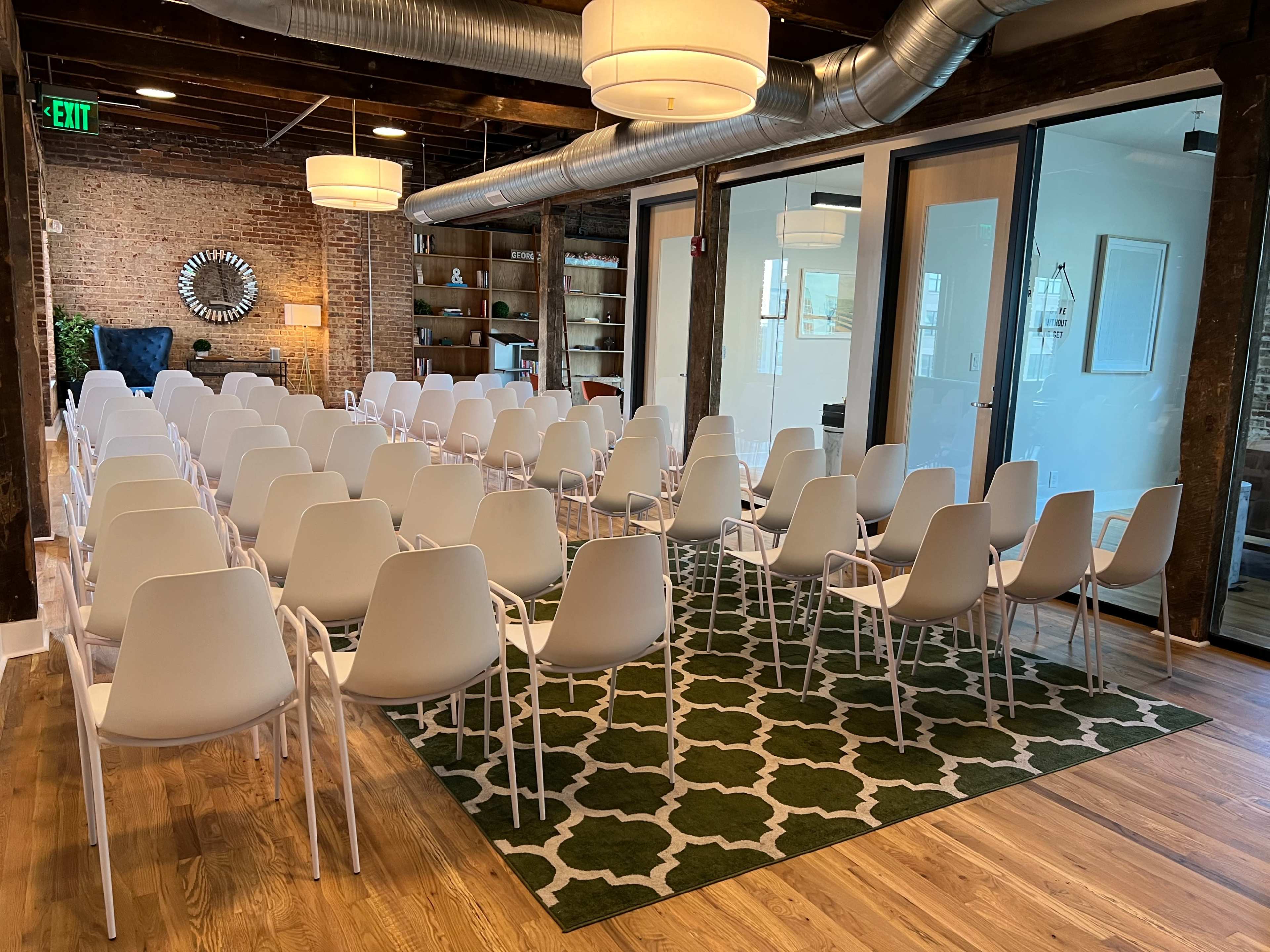 The image shows a spacious room set up with rows of white chairs on a green patterned rug, featuring exposed brick walls and a modern aesthetic.