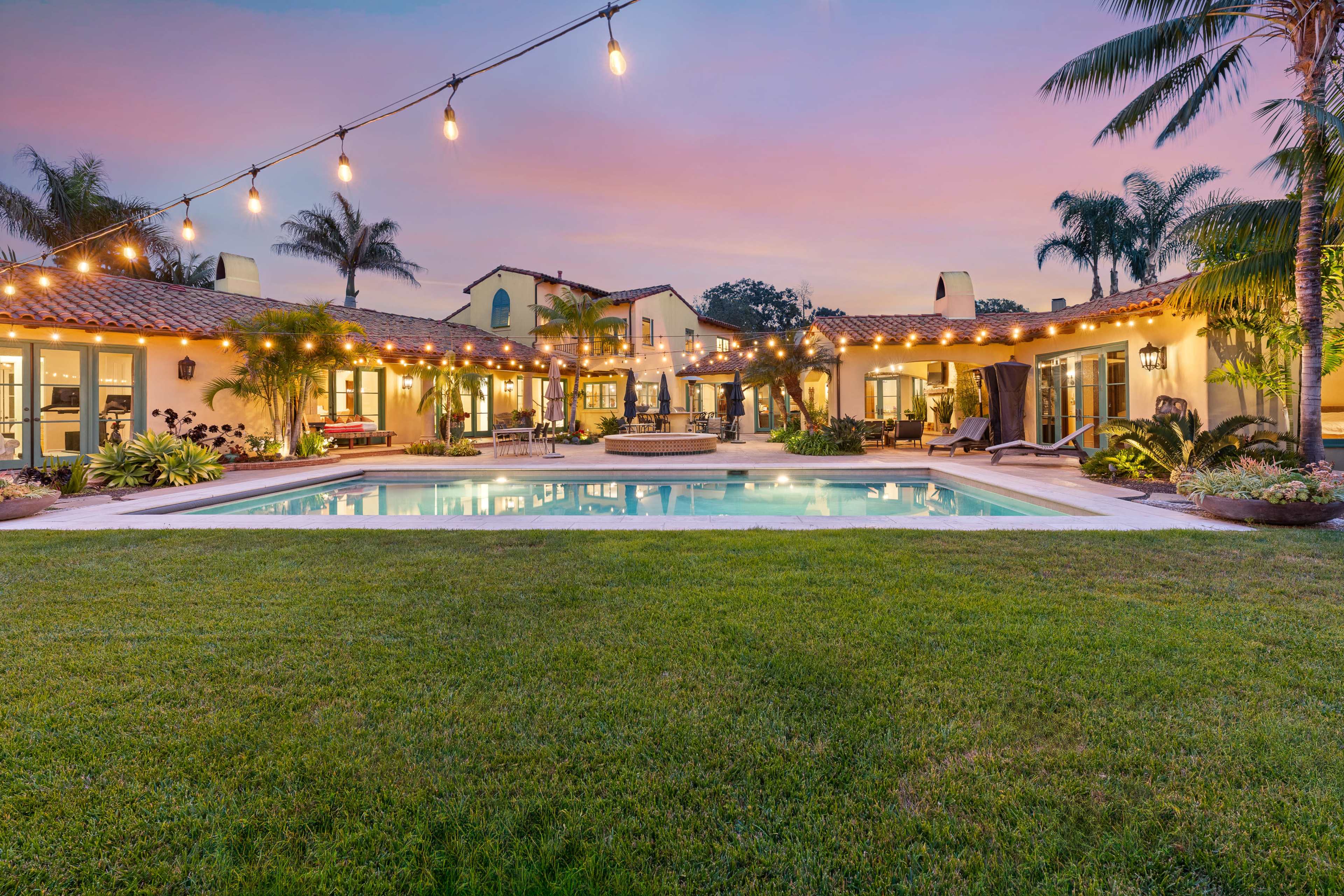 A spacious backyard features a swimming pool surrounded by lush grass and illuminated by string lights, with a Mediterranean-style house in the background.
