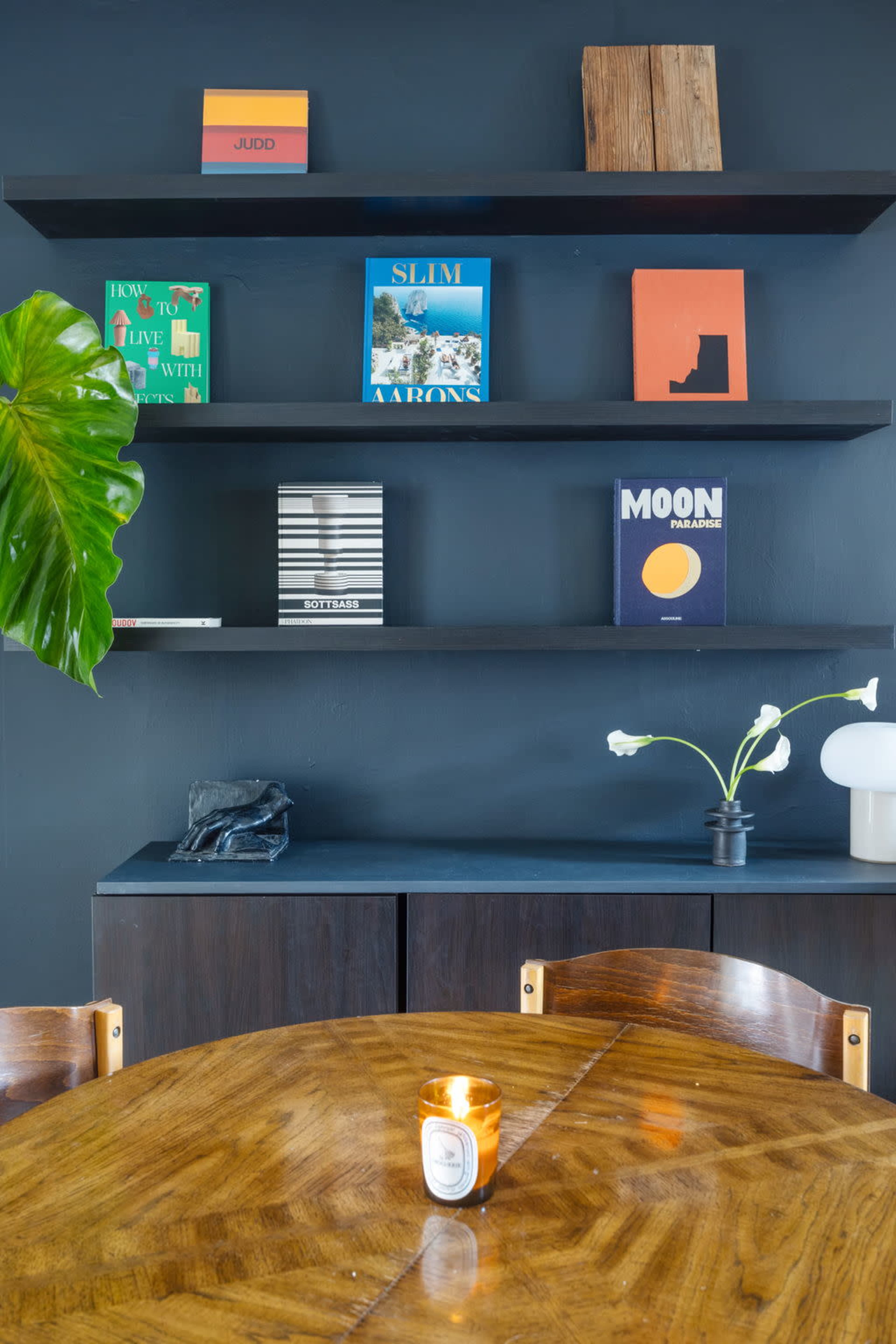 A wooden dining table is set in a room with dark walls, featuring shelves displaying colorful books and a decorative plant.