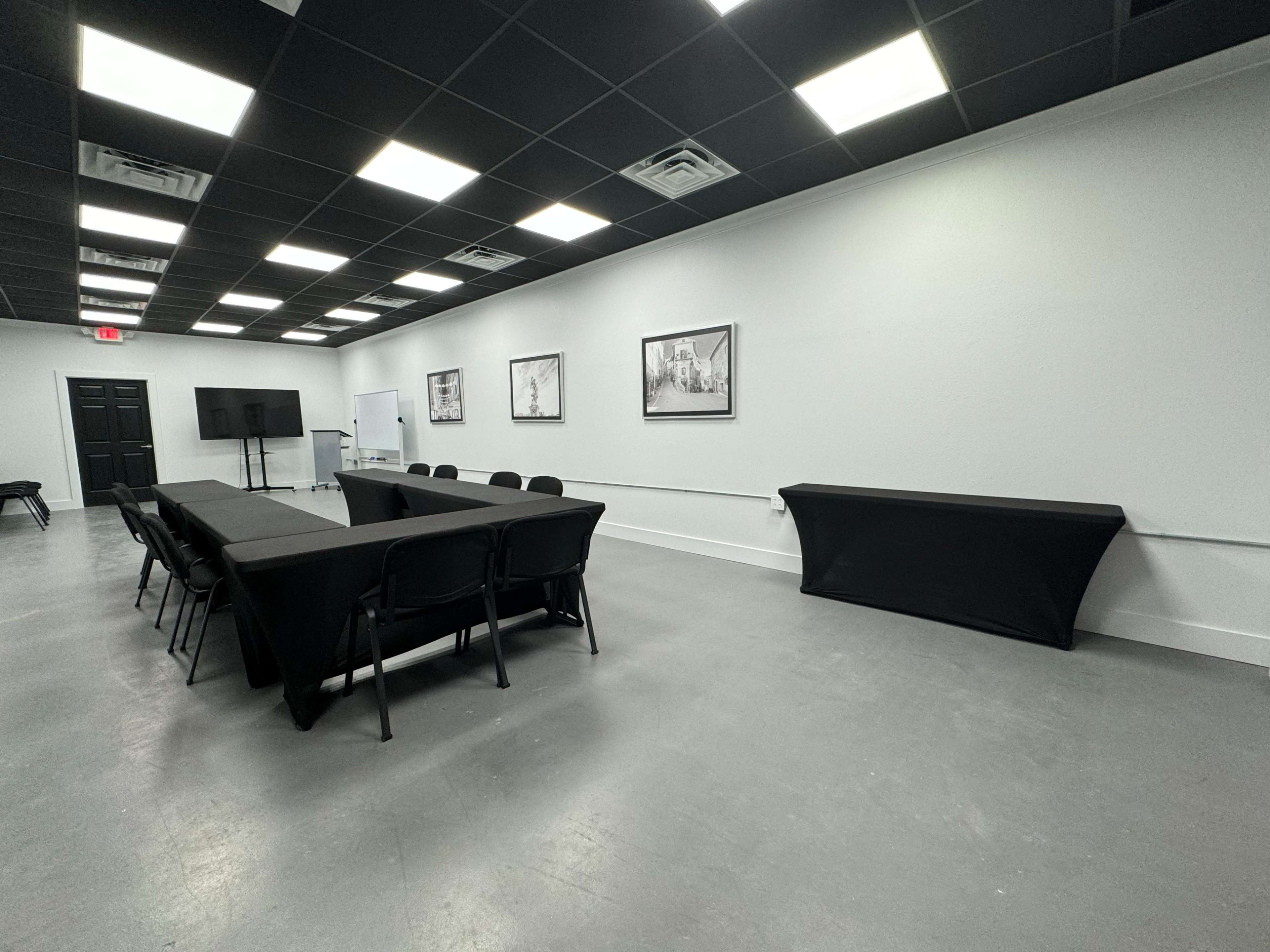 The image shows a modern meeting room with a long table and black chairs arranged in the center, flanked by white walls and ceiling lights.