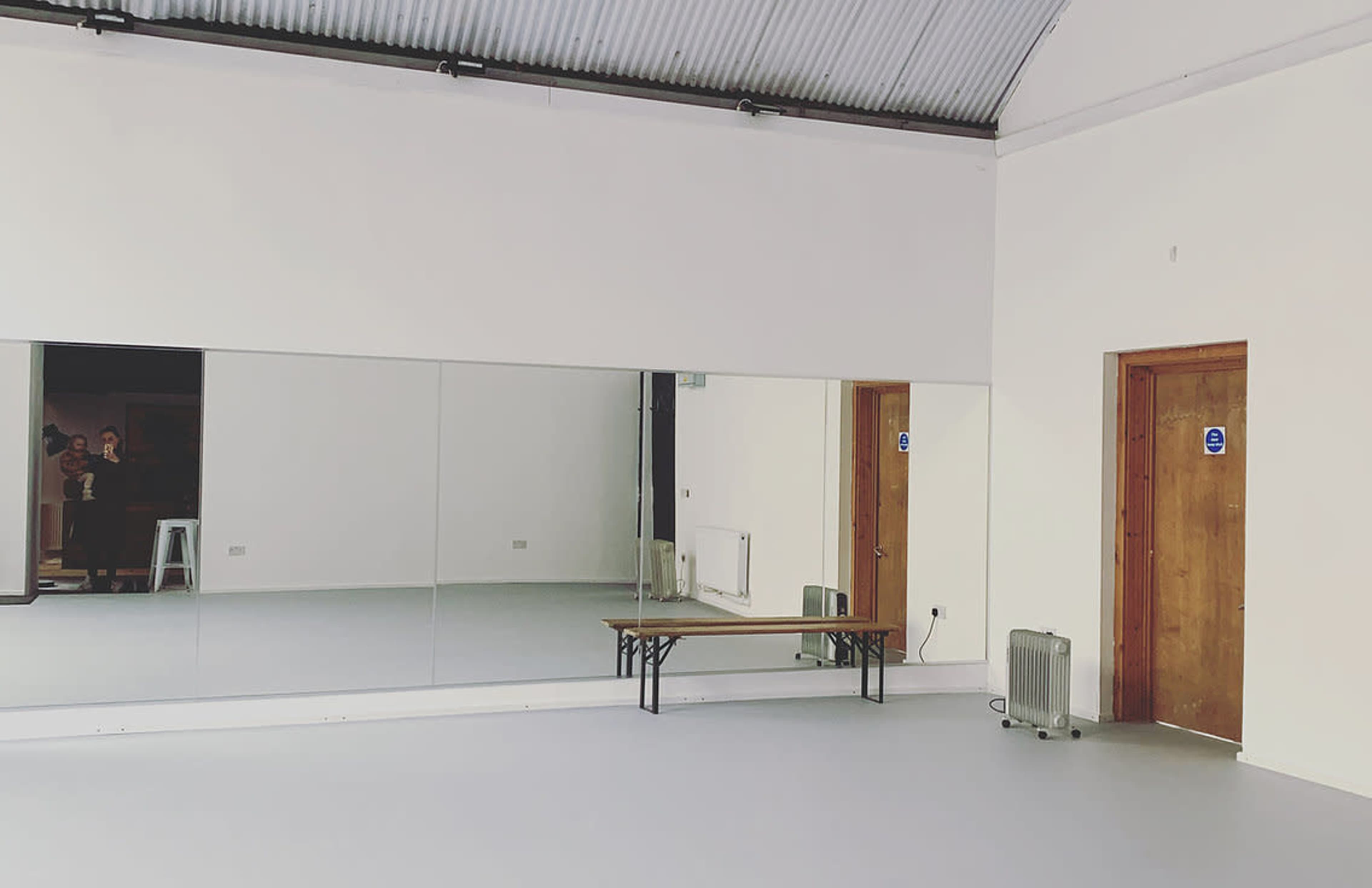 The image shows a spacious room with a large mirror, a wooden bench, a heater, and a door on one side.
