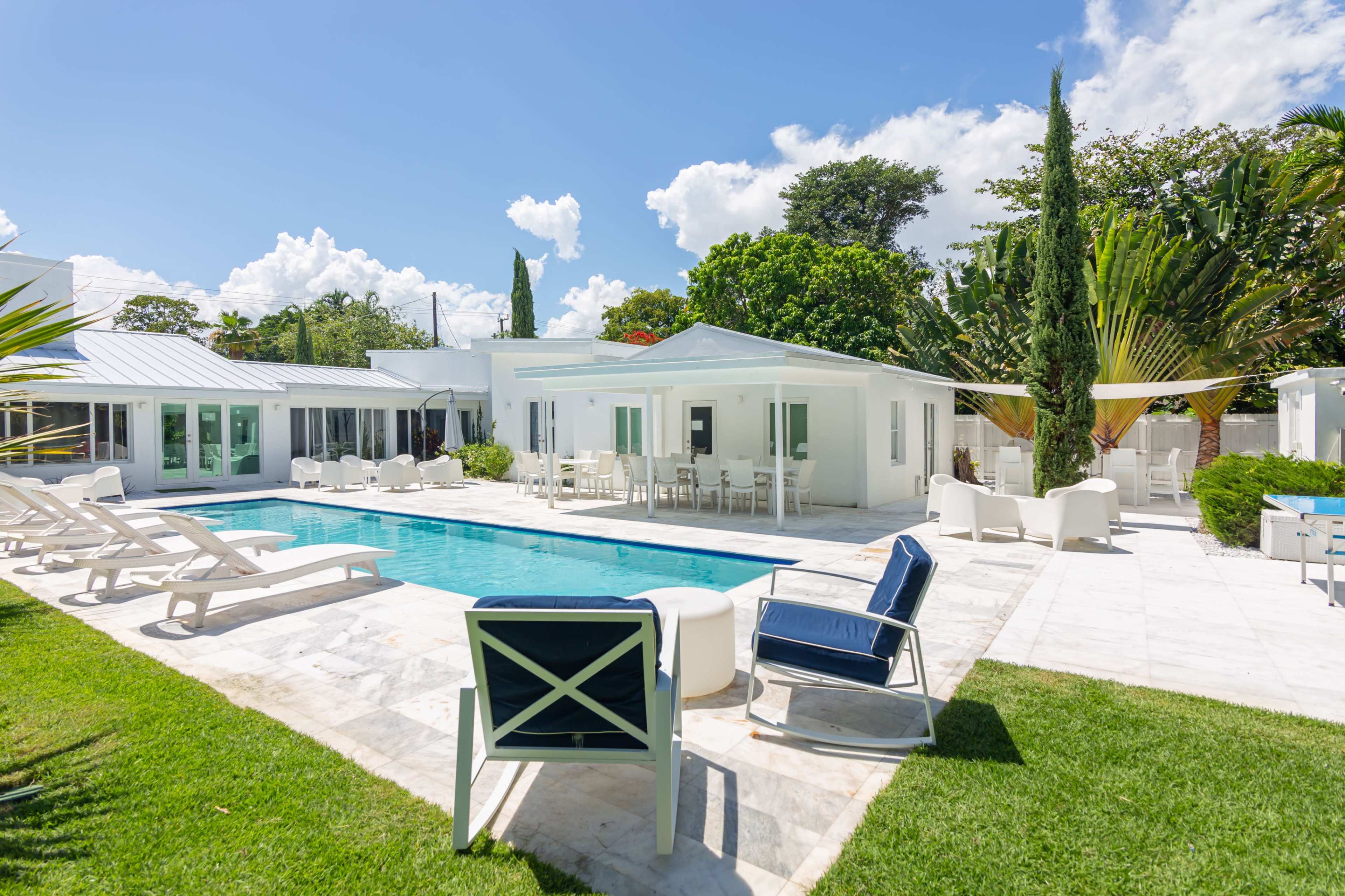 A modern backyard with a swimming pool, lounge chairs, and a white house surrounded by greenery and palm trees.
