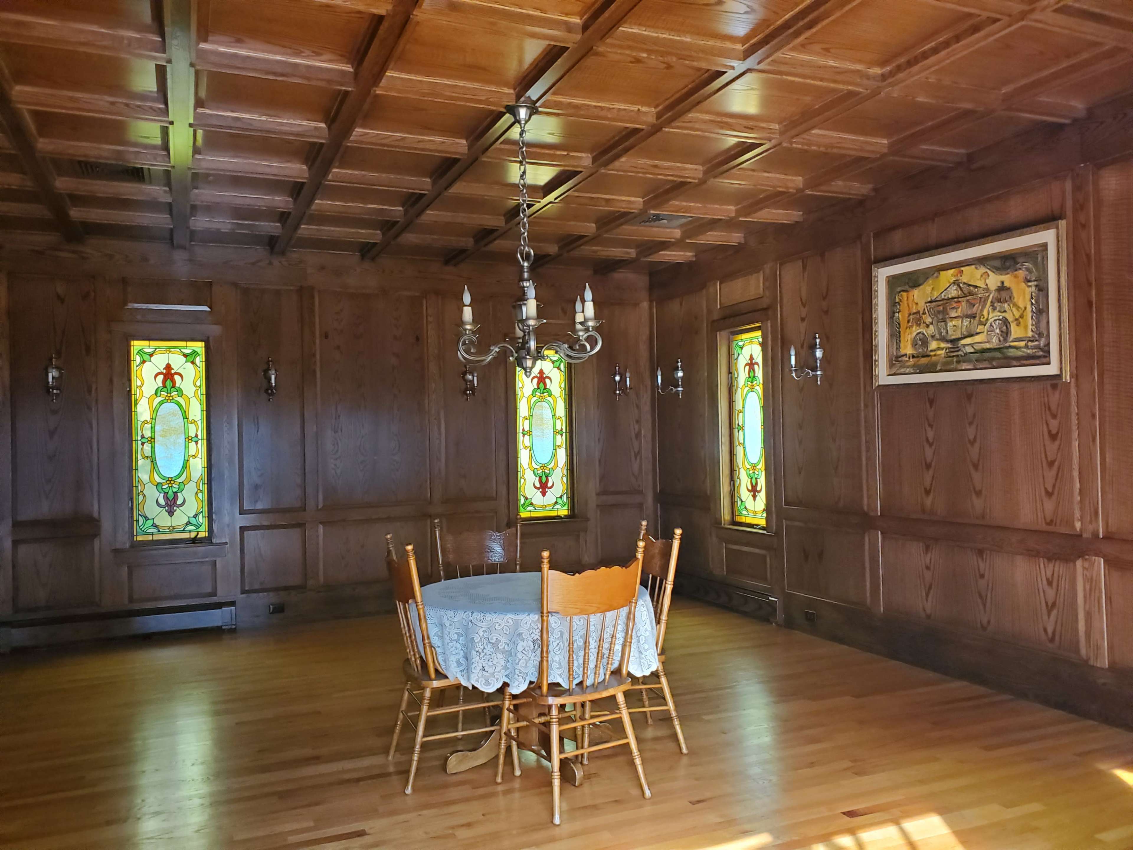 A wooden-paneled dining room features a round table surrounded by chairs, with stained glass windows illuminating the space.