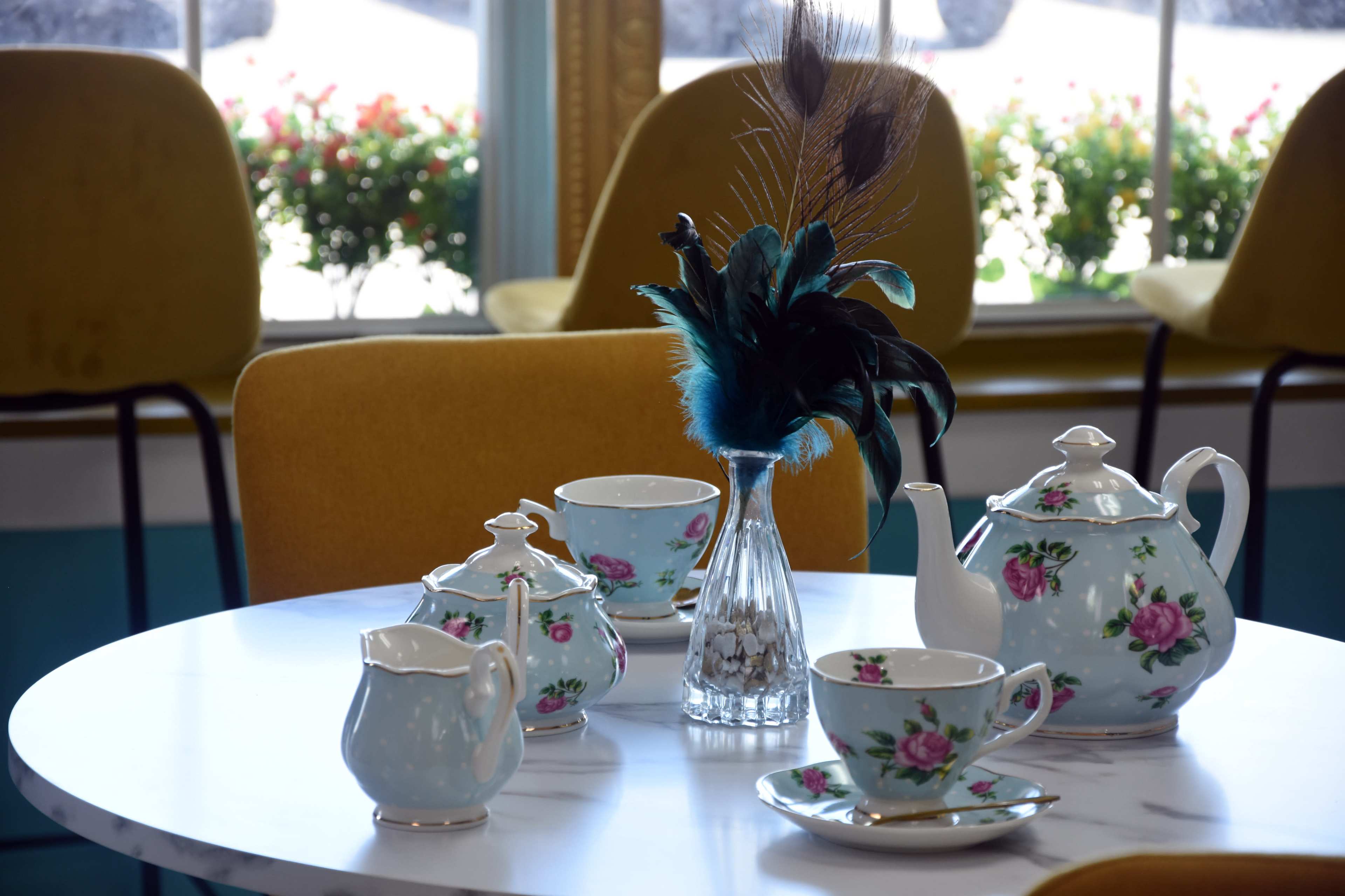 A vintage tea set with floral patterns is arranged on a marble table next to a decorative vase with peacock feathers, while chairs are positioned around the table in a brightly lit room.