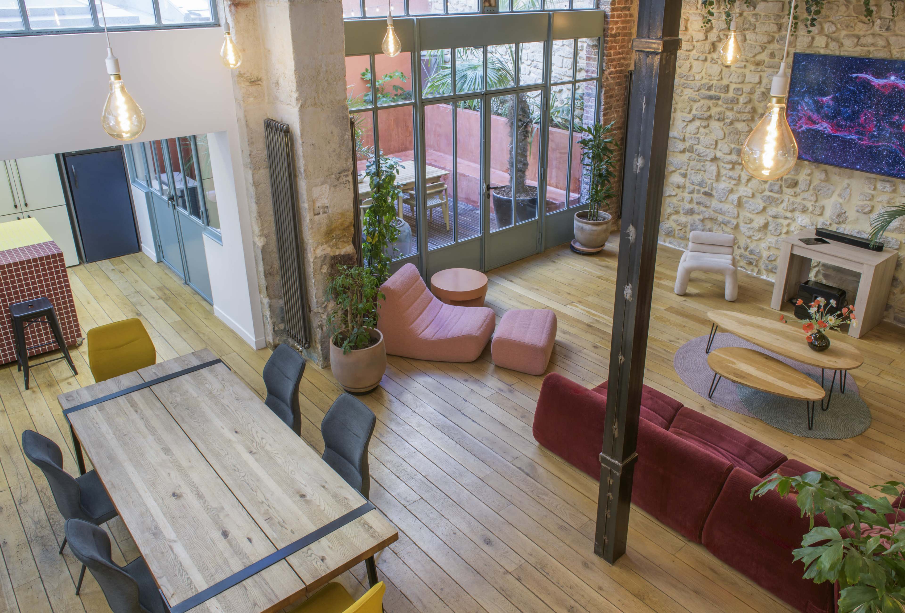 The image shows a spacious, modern open-plan office with large windows, wooden floors, a dining table, colorful seating areas, and plants.