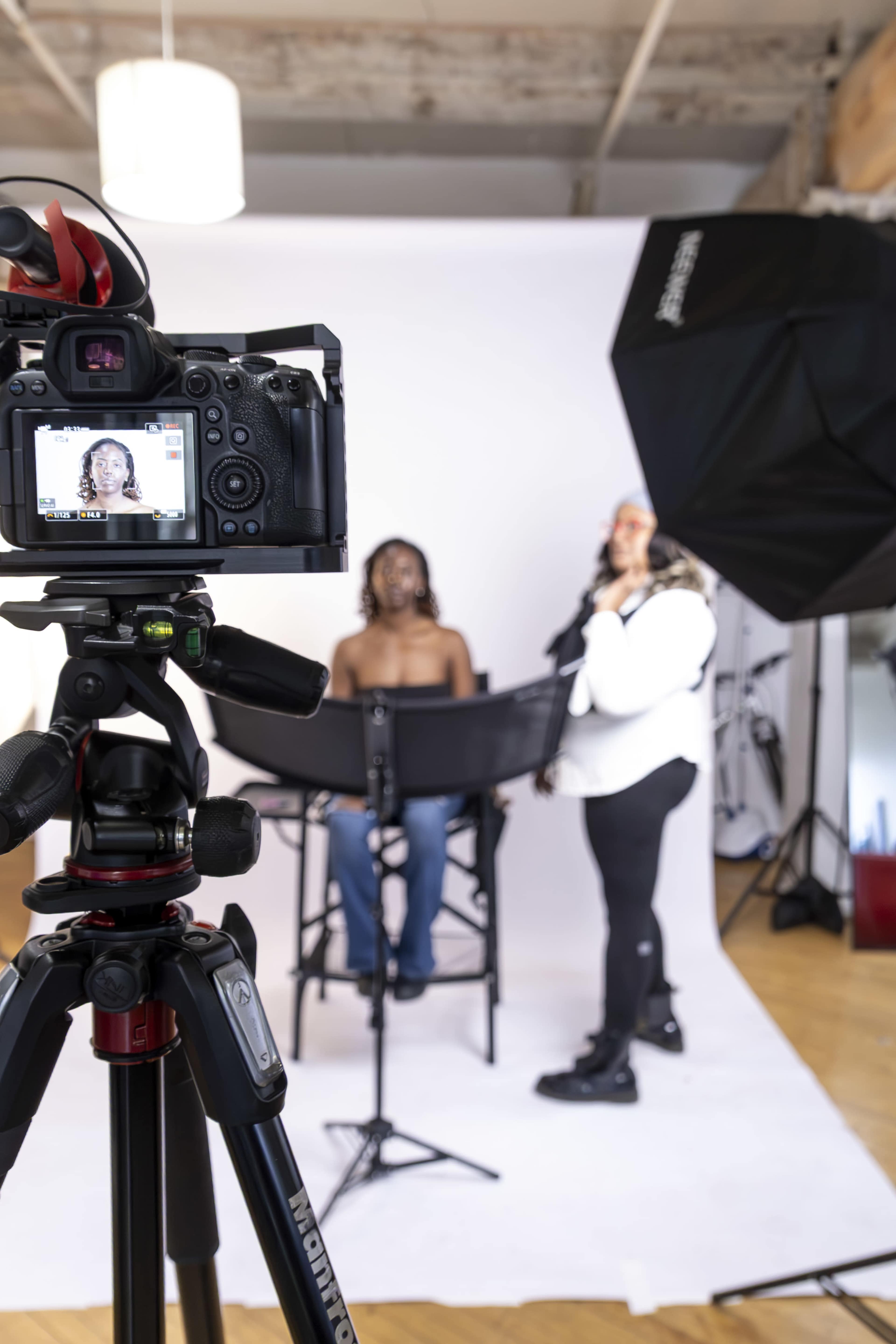 A photograph captures a camera set on a tripod focusing on a person seated in a studio, while another individual stands nearby holding a makeup brush.