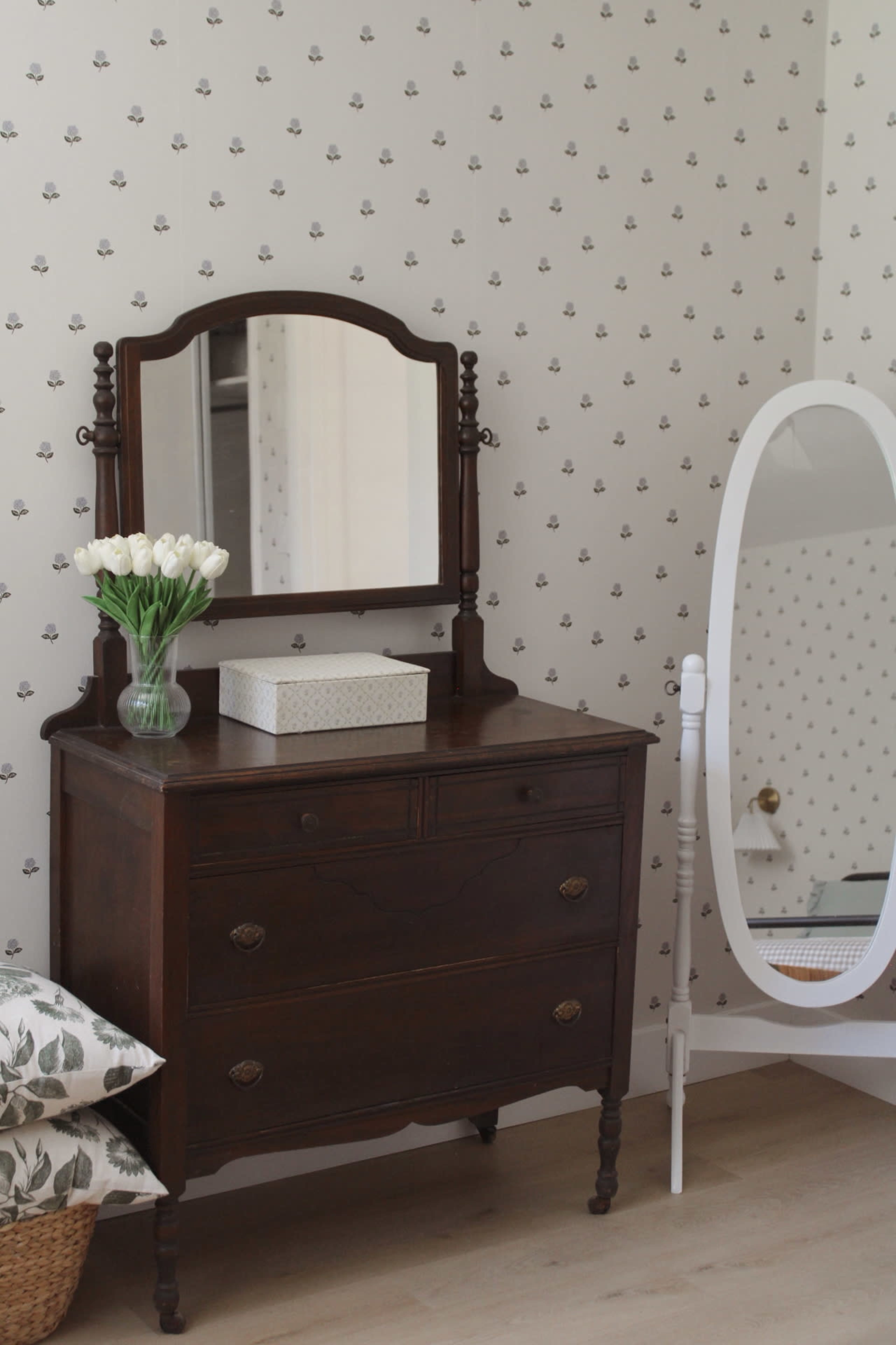 A wooden dresser with a mirror is placed against a wallpapered wall, alongside a white oval mirror and a vase of white flowers.