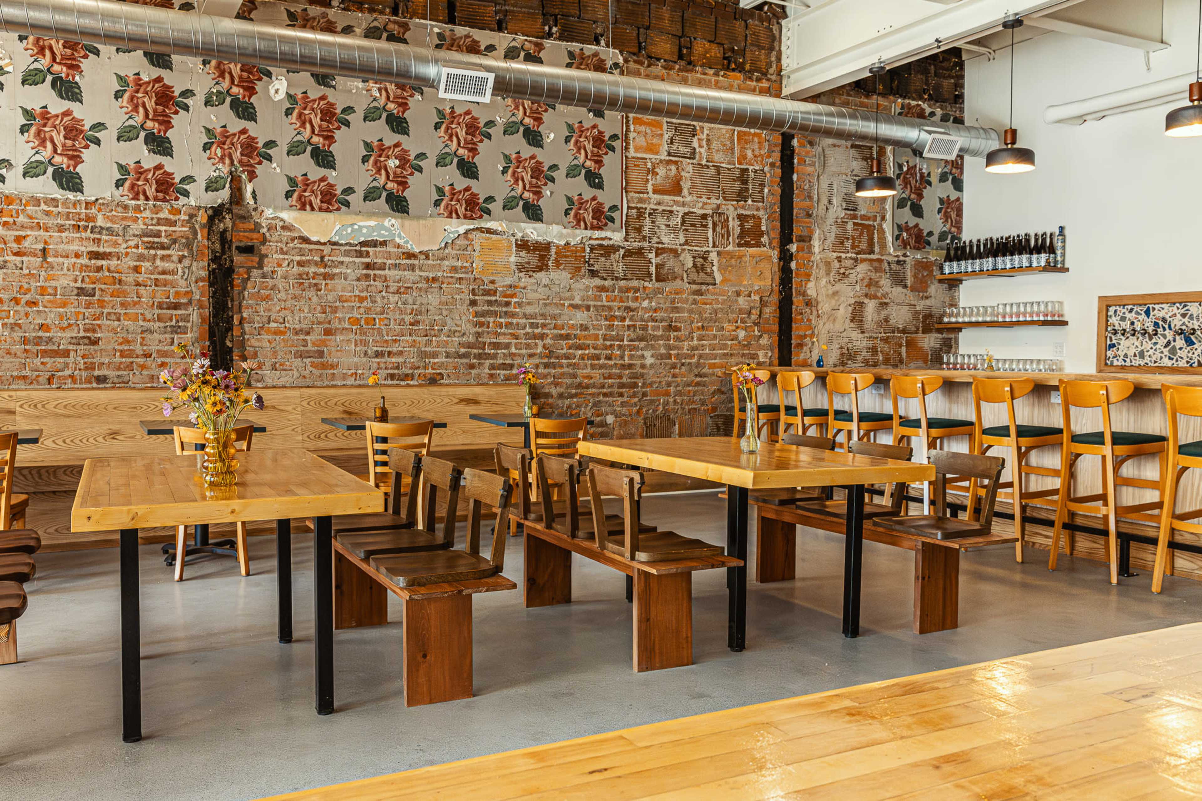 The image shows a spacious restaurant interior with wooden tables and benches, brick walls adorned with floral wallpaper, and a bar area featuring high stools.