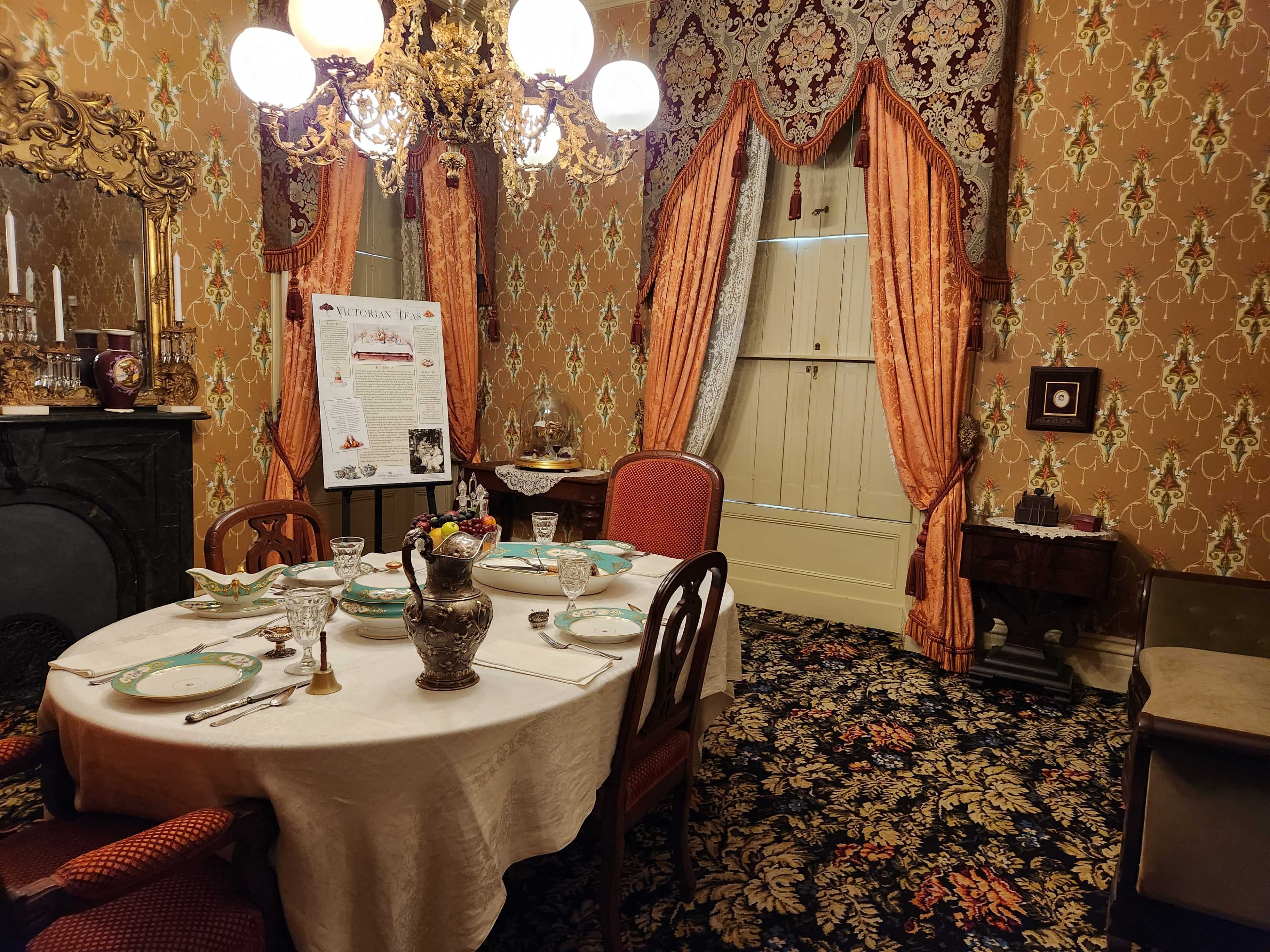 A formal dining room set with an ornate table, decorative chairs, and patterned wallpaper, featuring a chandelier and a display of elegant tableware.