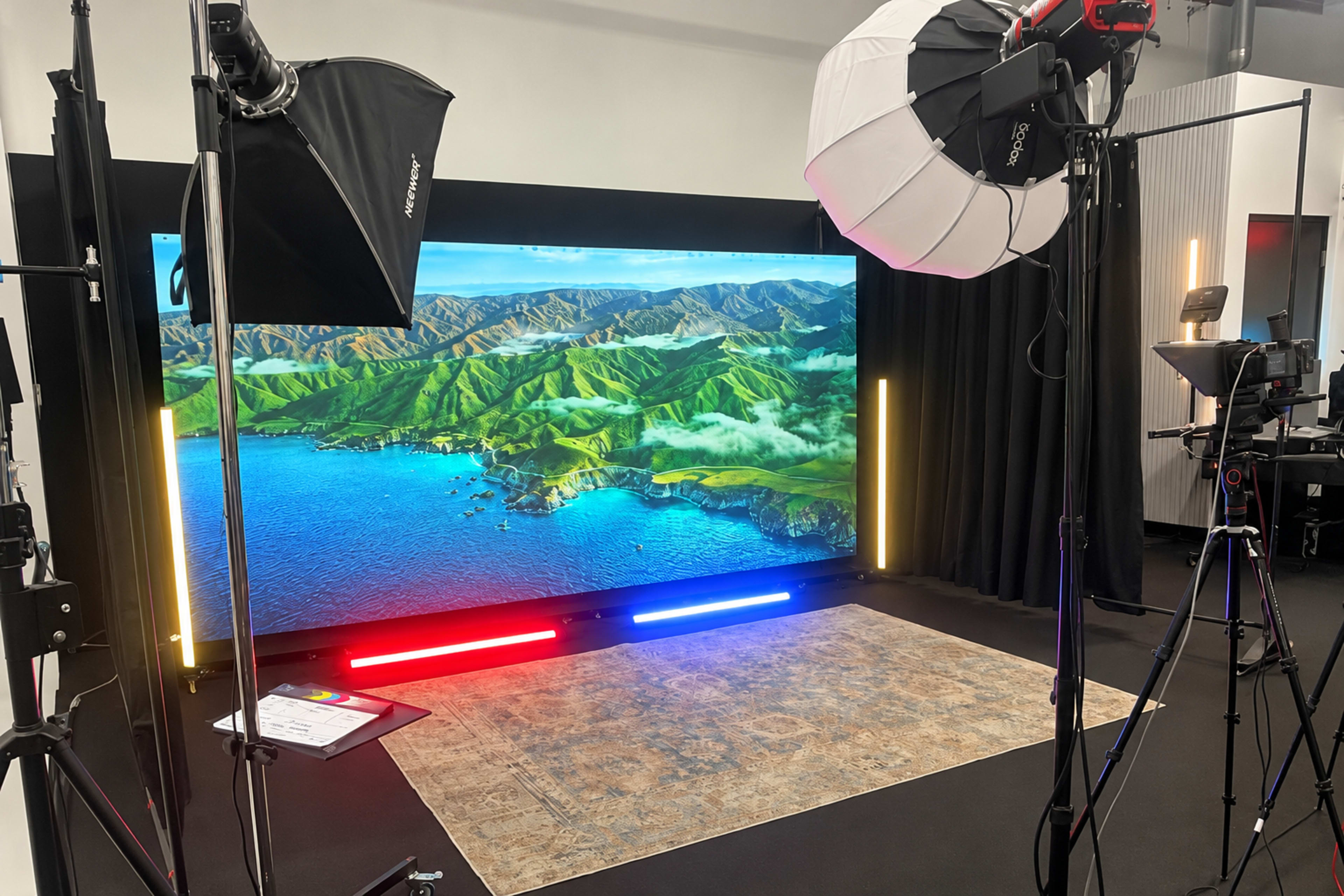 The image shows a photography studio setup with a large screen displaying a coastal landscape, flanked by studio lights and camera equipment.